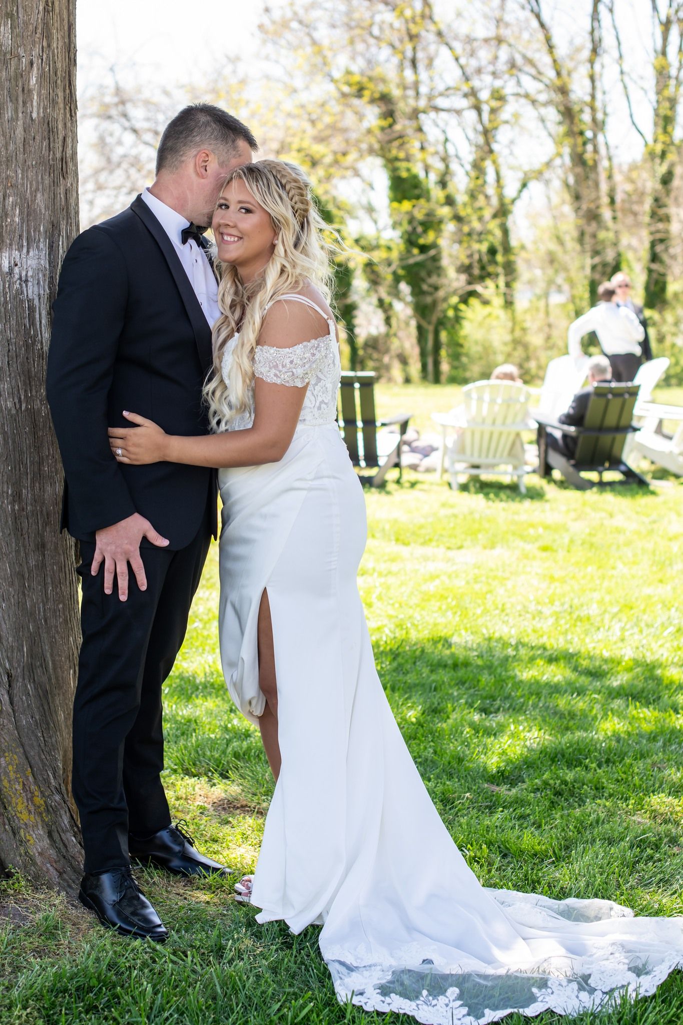 Bride and groom share a romantic moment leaning against a tree outdoors on their wedding day.