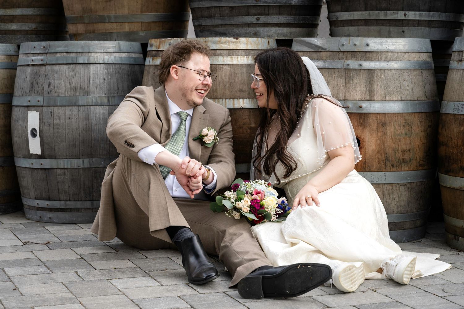 Bride and groom sitting together on cobblestones, holding a colorful bouquet, smiling at each other in front of rustic wine barrels.