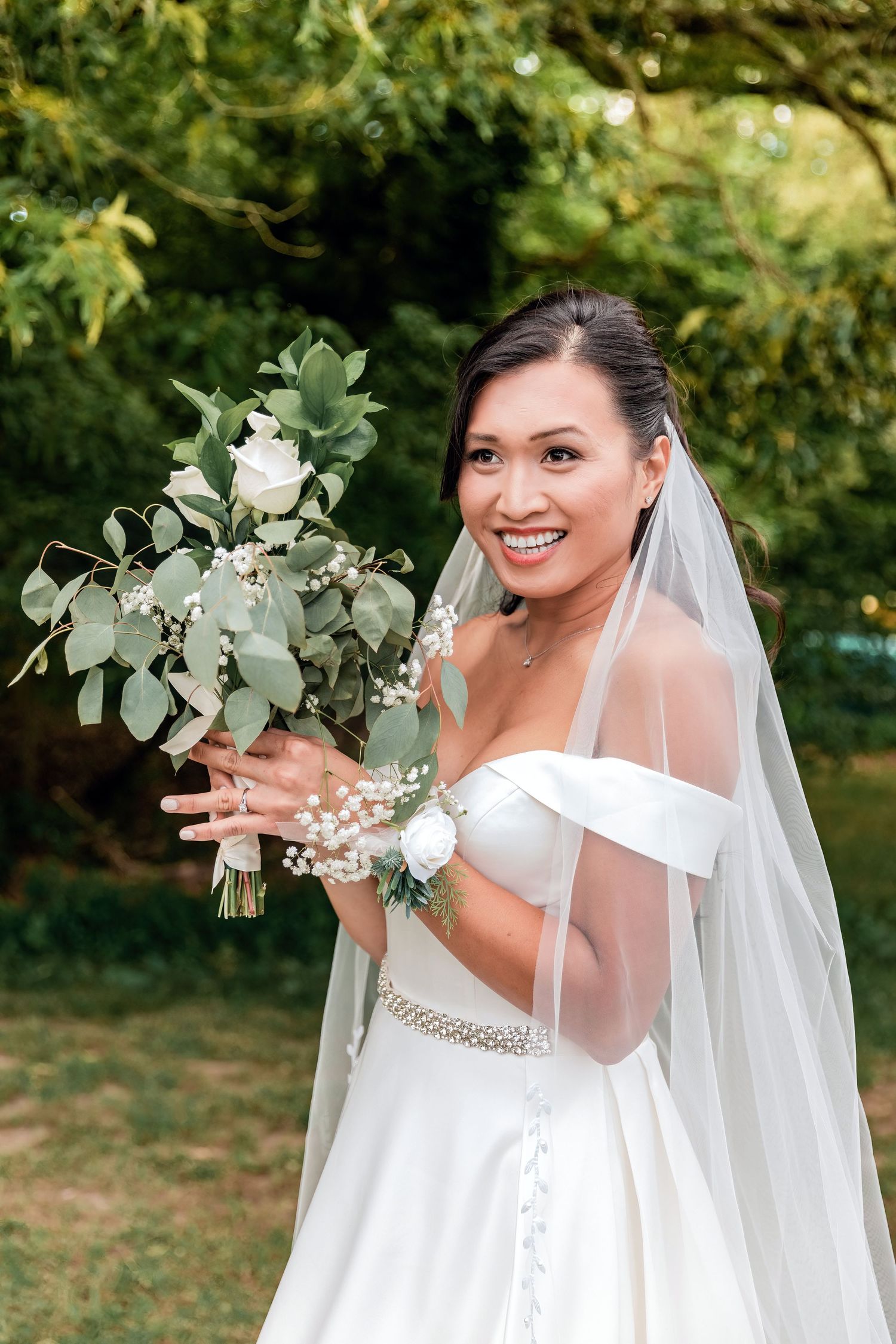 Smiling Asian bride in white off-shoulder gown and veil holds green and white bouquet outdoors.