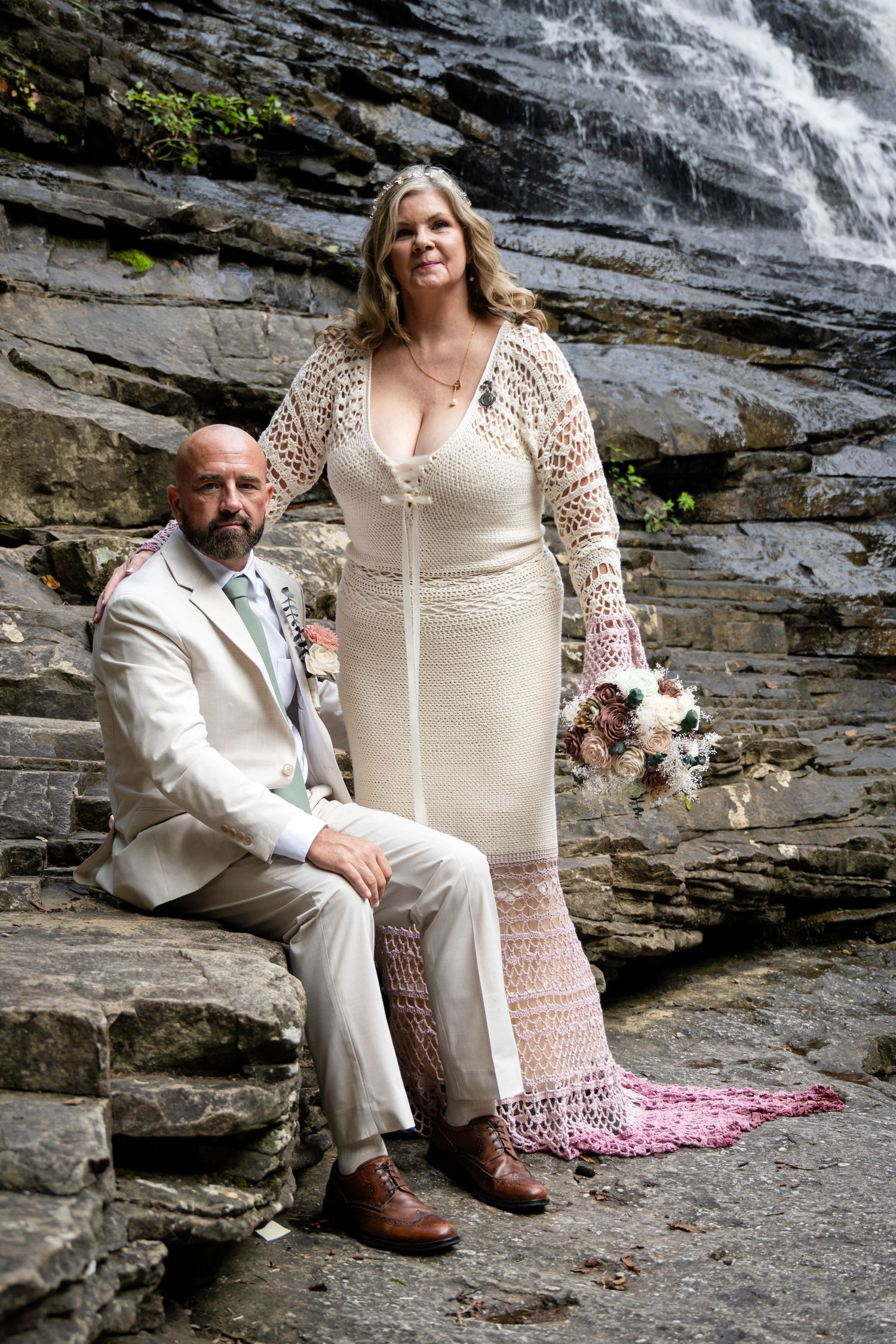 Bride in crochet gown and groom in tan suit pose on rocky steps before a waterfall for their wedding portrait.