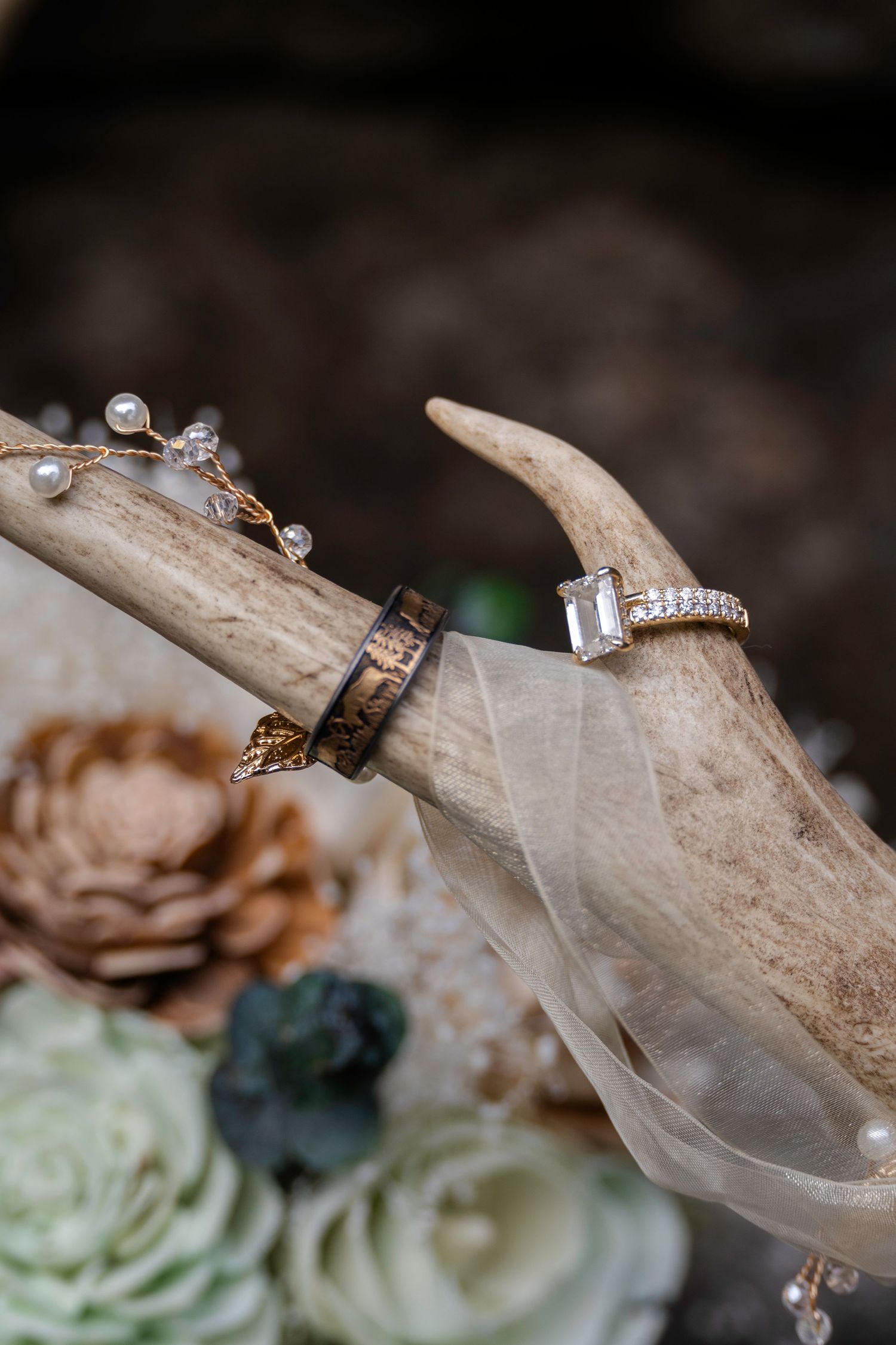 Wedding rings displayed on deer antler with diamond engagement ring, dark band, and pearl hair accessory with floral decor.