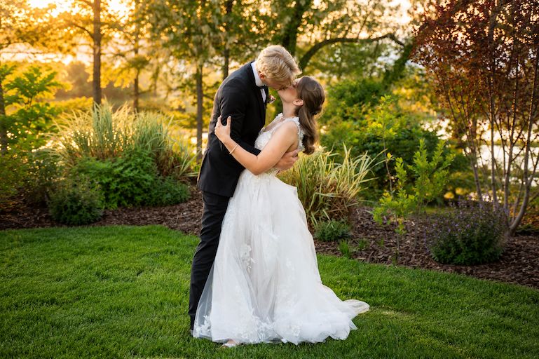 Bride and groom share a romantic dip kiss at golden hour in a lush garden setting.