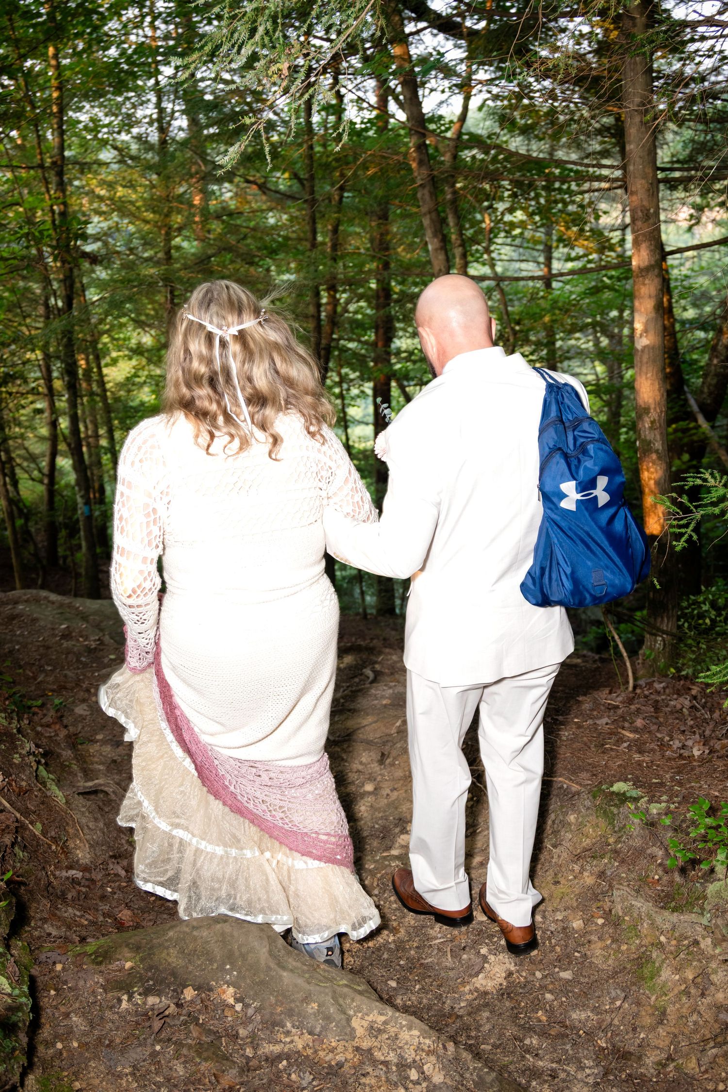 Couple in white wedding attire walking hand-in-hand through a lush forest, man carrying blue Under Armour backpack.