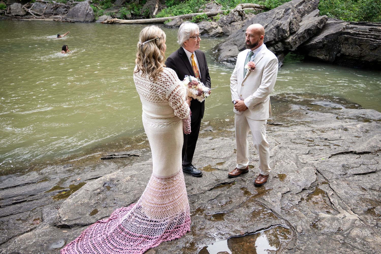 Outdoor wedding ceremony on rocky riverbank with bride in lace gown, groom in white suit, and officiant.