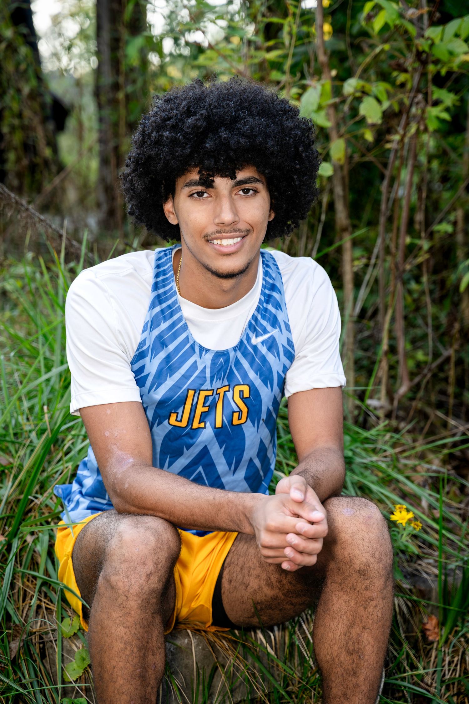 Young man in Jets jersey and gold shorts sits outdoors smiling, with curly hair and green foliage background.