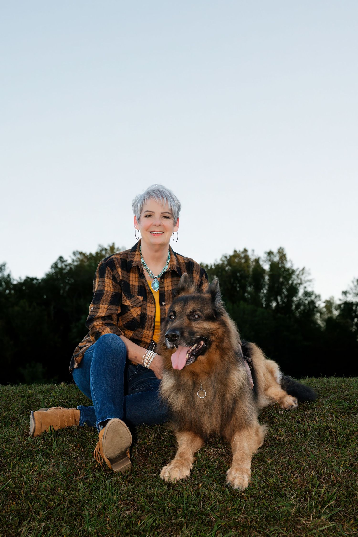 Woman with short gray hair in plaid shirt sits on grass with a fluffy German Shepherd dog outdoors.