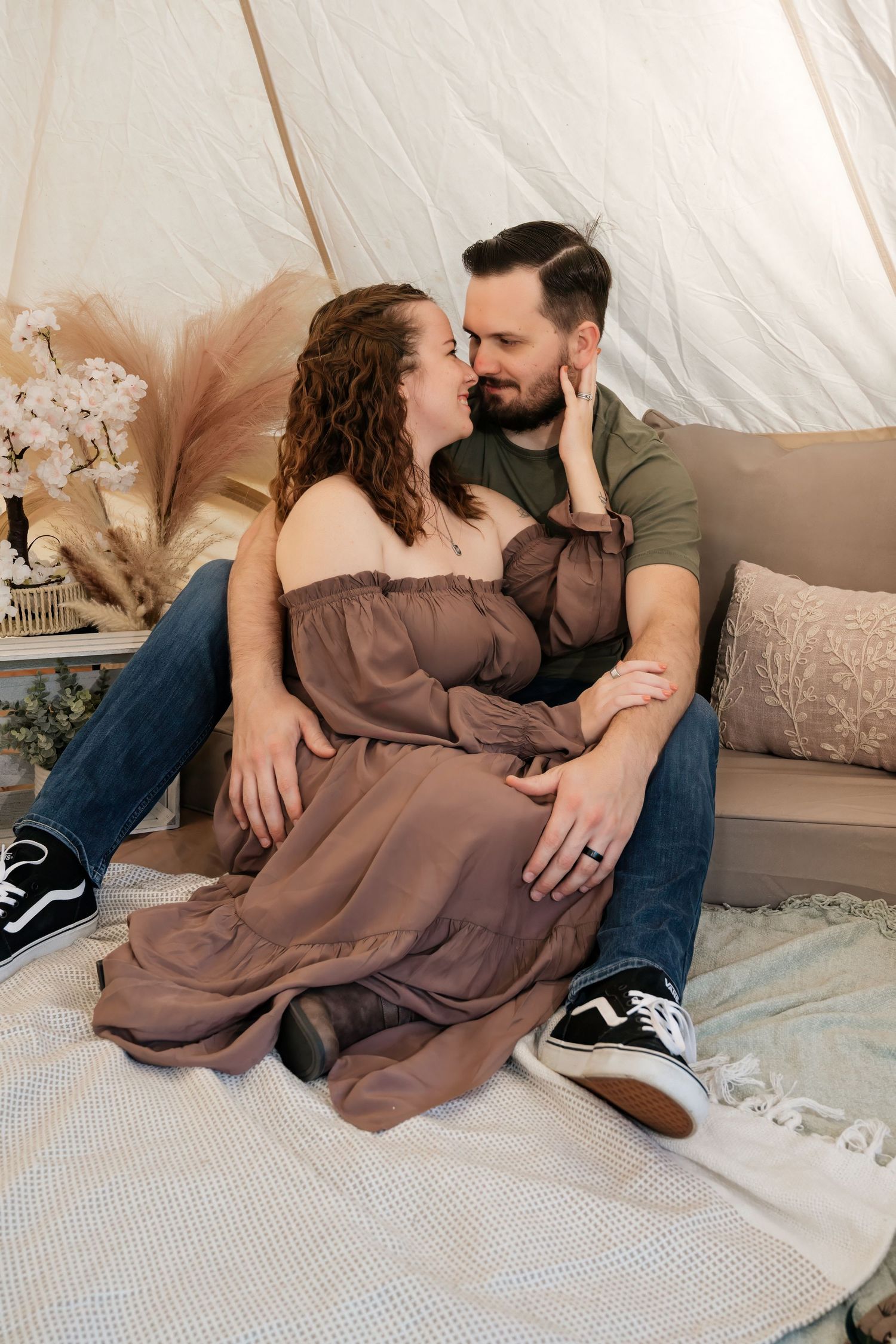 A couple shares a romantic moment sitting together in a boho tent, the woman in a brown dress touching the man's face.