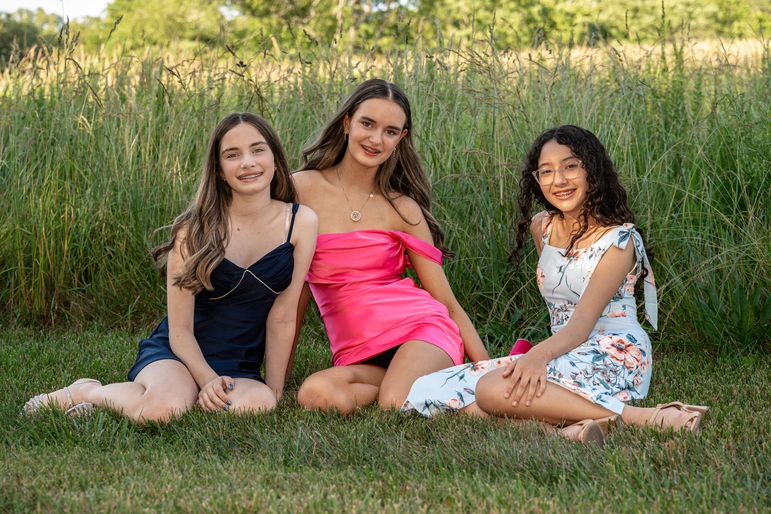 Three teenage girls in colorful summer dresses sitting together on green grass with tall reeds in the background.