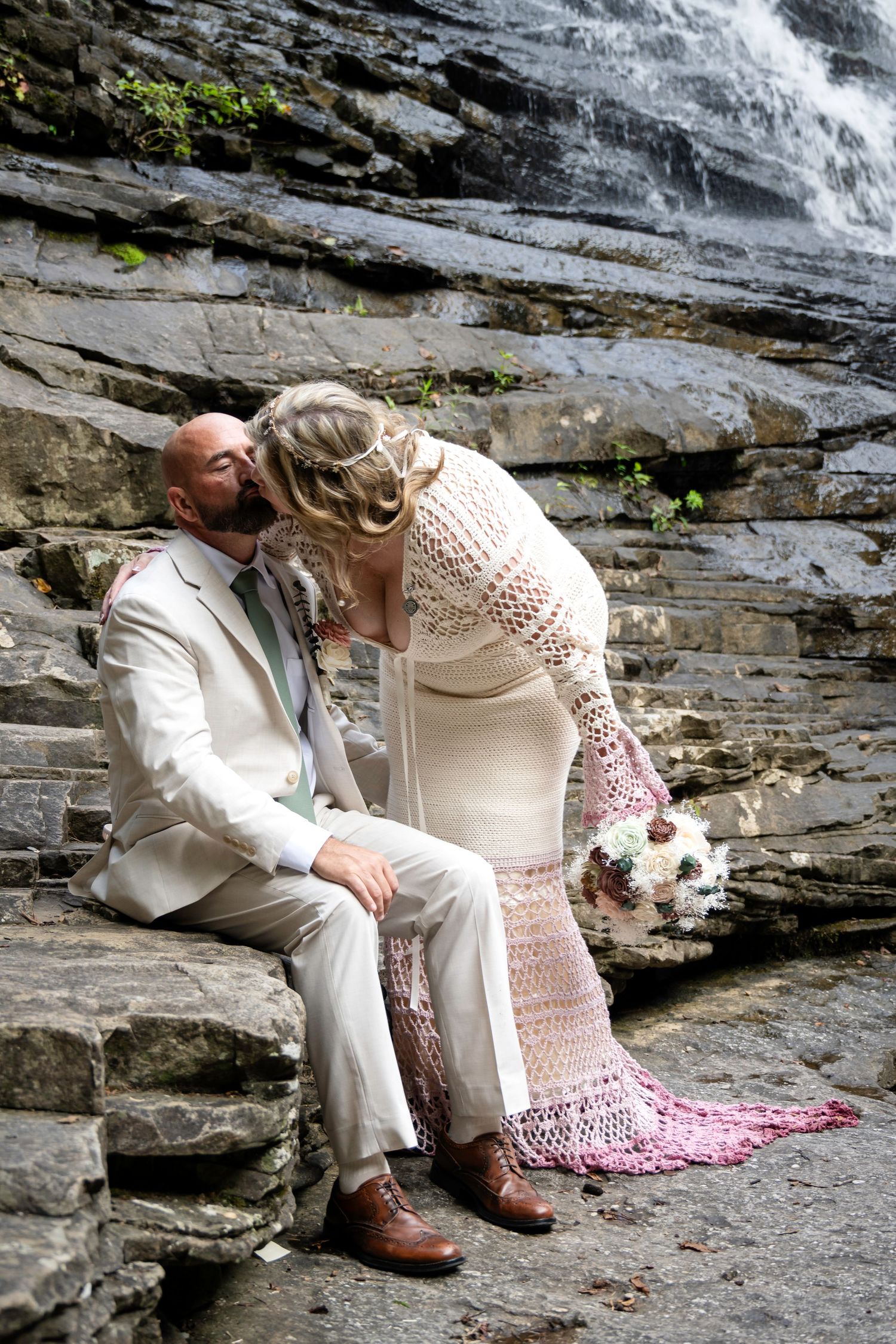 Bride and groom share a kiss on stone steps before a waterfall, bride in lace dress holding bouquet, groom in white suit.