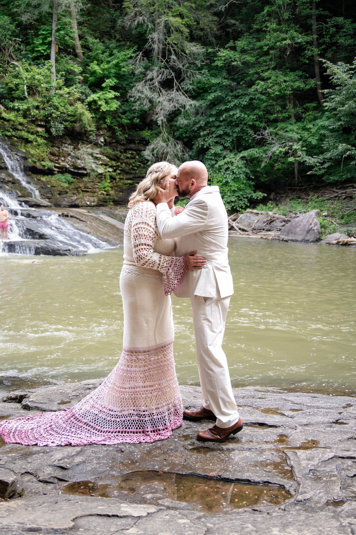 Bride and groom share a kiss in front of a waterfall, bride wearing a white crochet dress with pink hem.