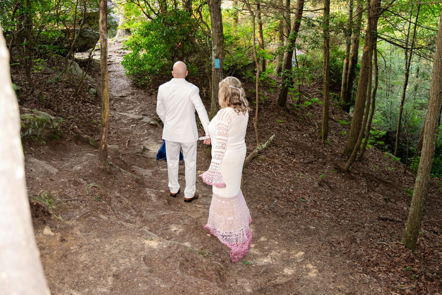A couple in formal white attire walks hand-in-hand along a wooded forest path during their wedding day.