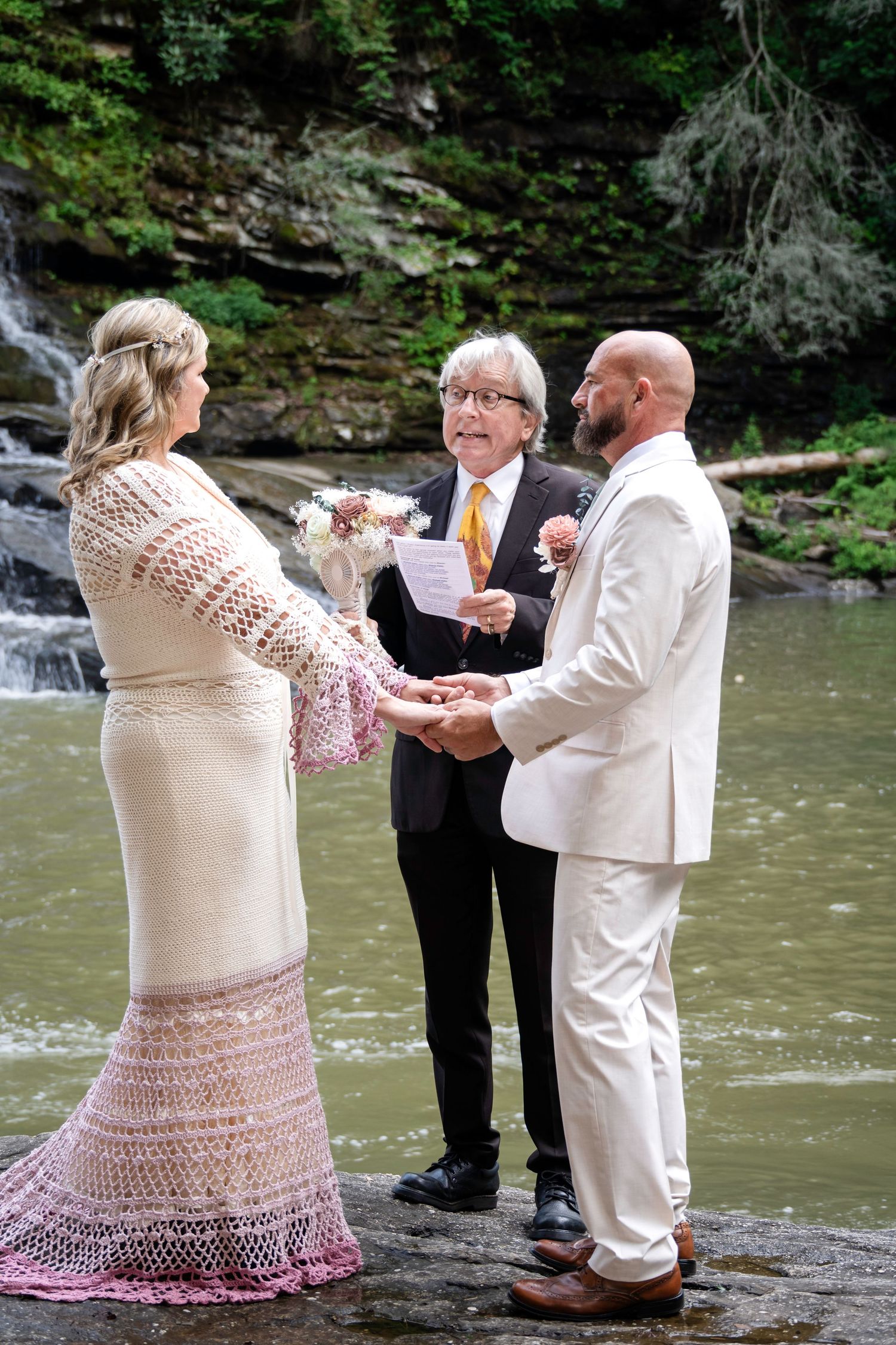 Couple exchanges vows with officiant by a waterfall, bride in lace dress, groom in white suit.