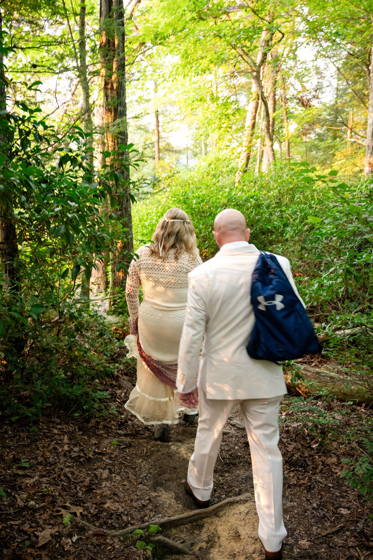 Couple in wedding attire walking hand-in-hand along a forest path surrounded by lush green trees.