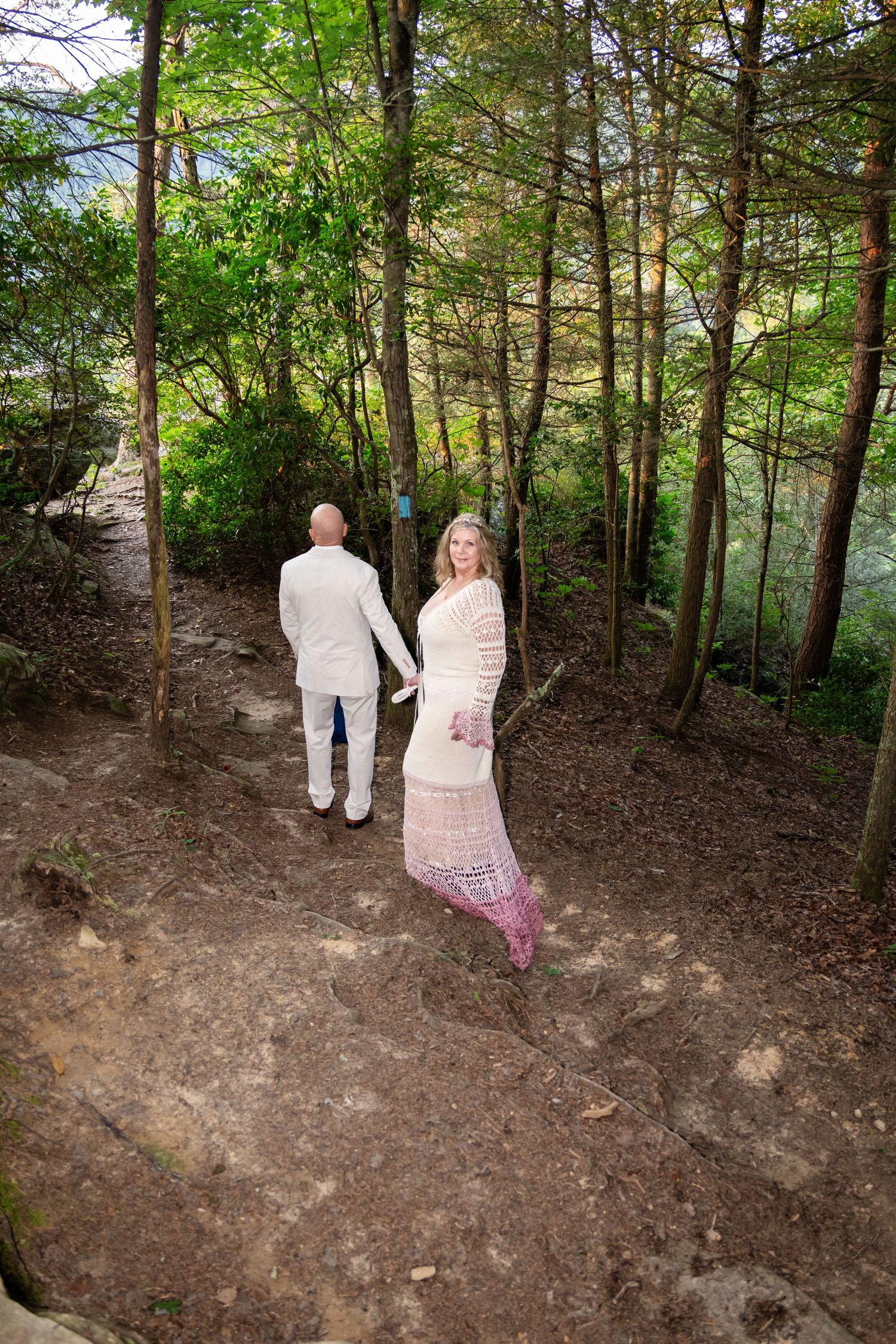Couple in white attire holding hands on a forest trail, woman in lace dress looks back at camera.