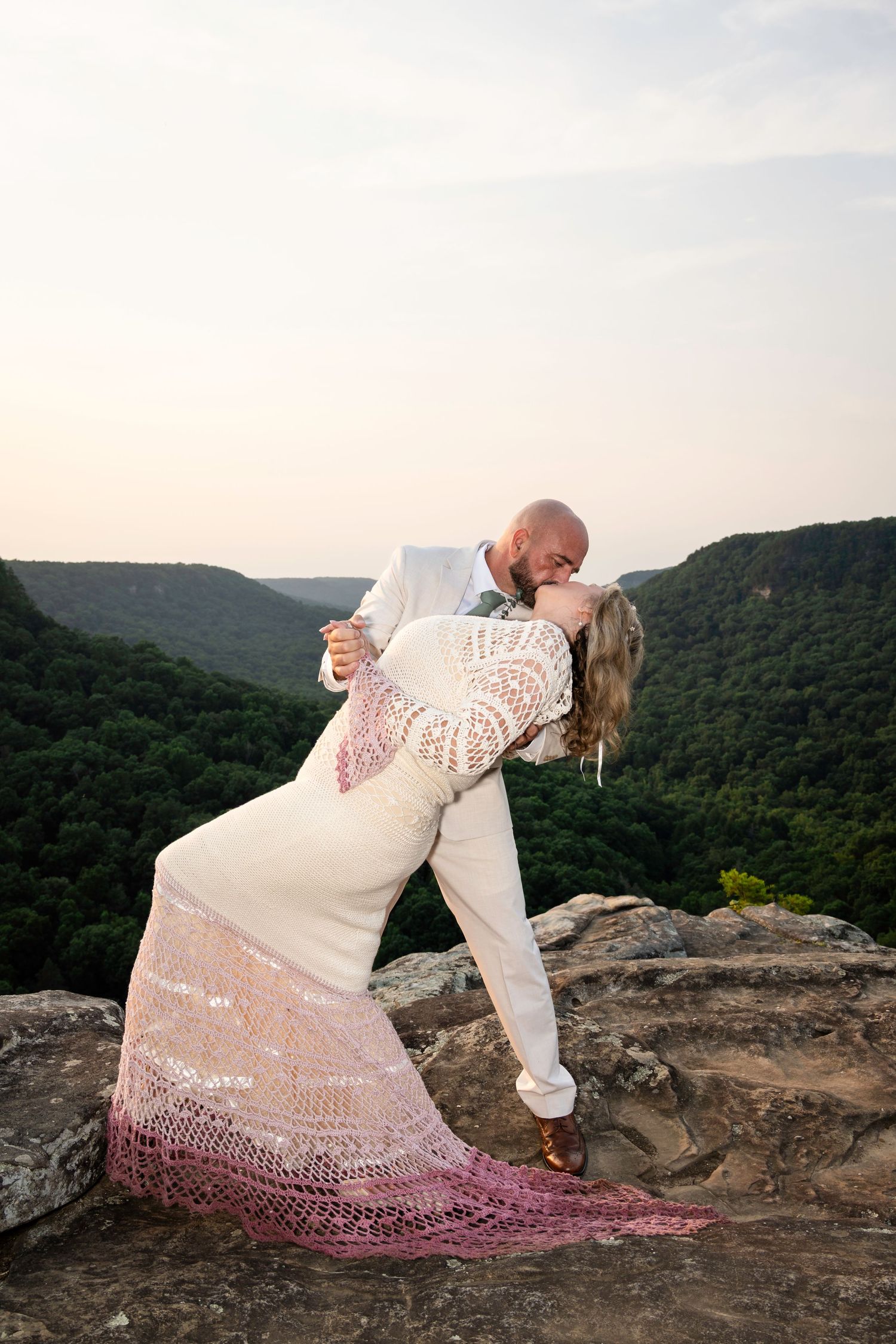 Bride and groom share a romantic dip kiss on a rocky mountain overlook with lush green valleys at sunset.