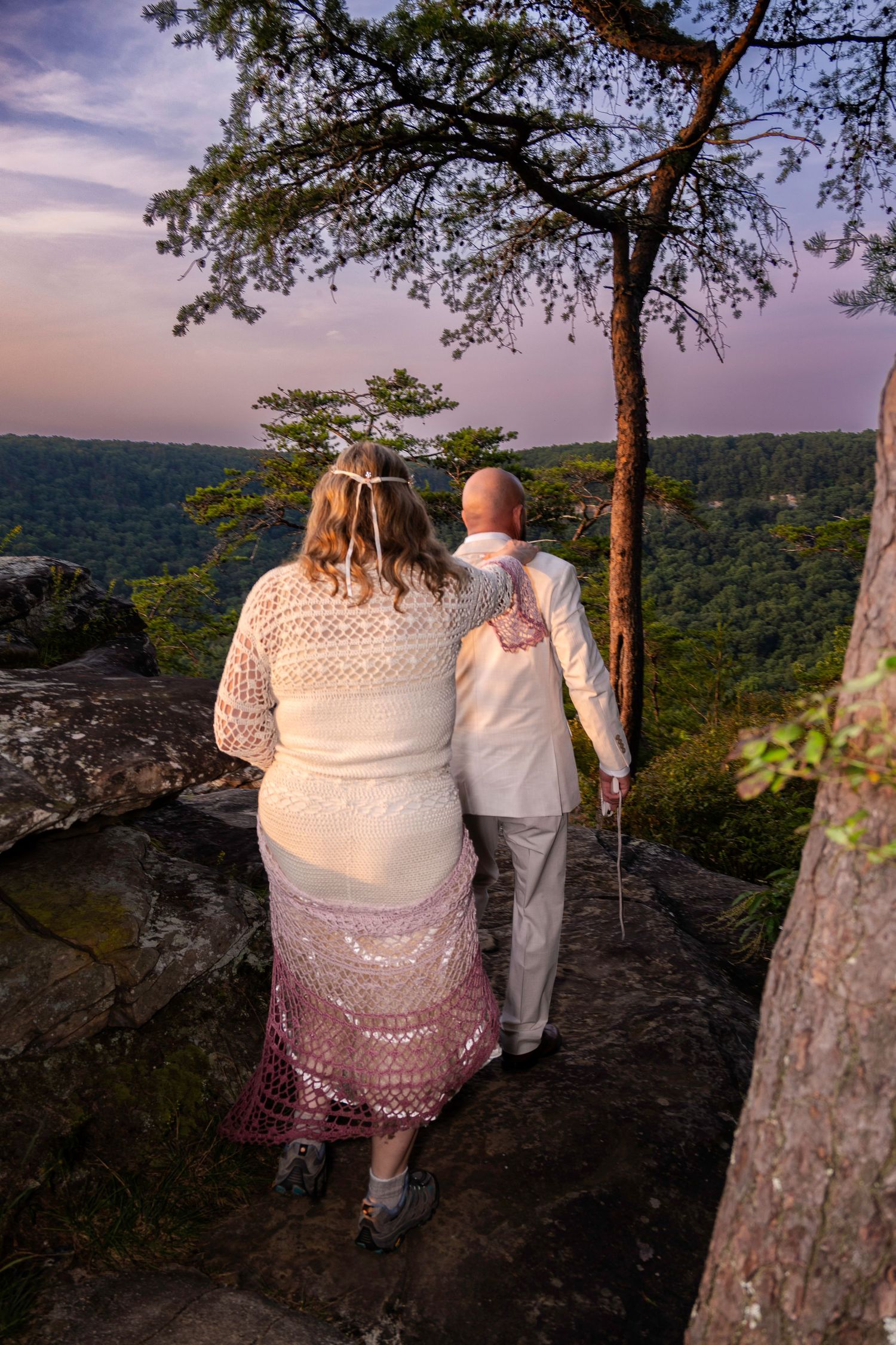 Couple in wedding attire standing on rocky overlook at sunset, surrounded by lush green forest and purple sky.