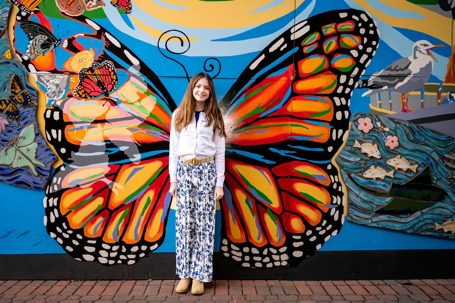 Woman posing in front of colorful butterfly mural wall art, wearing white top and floral pants.