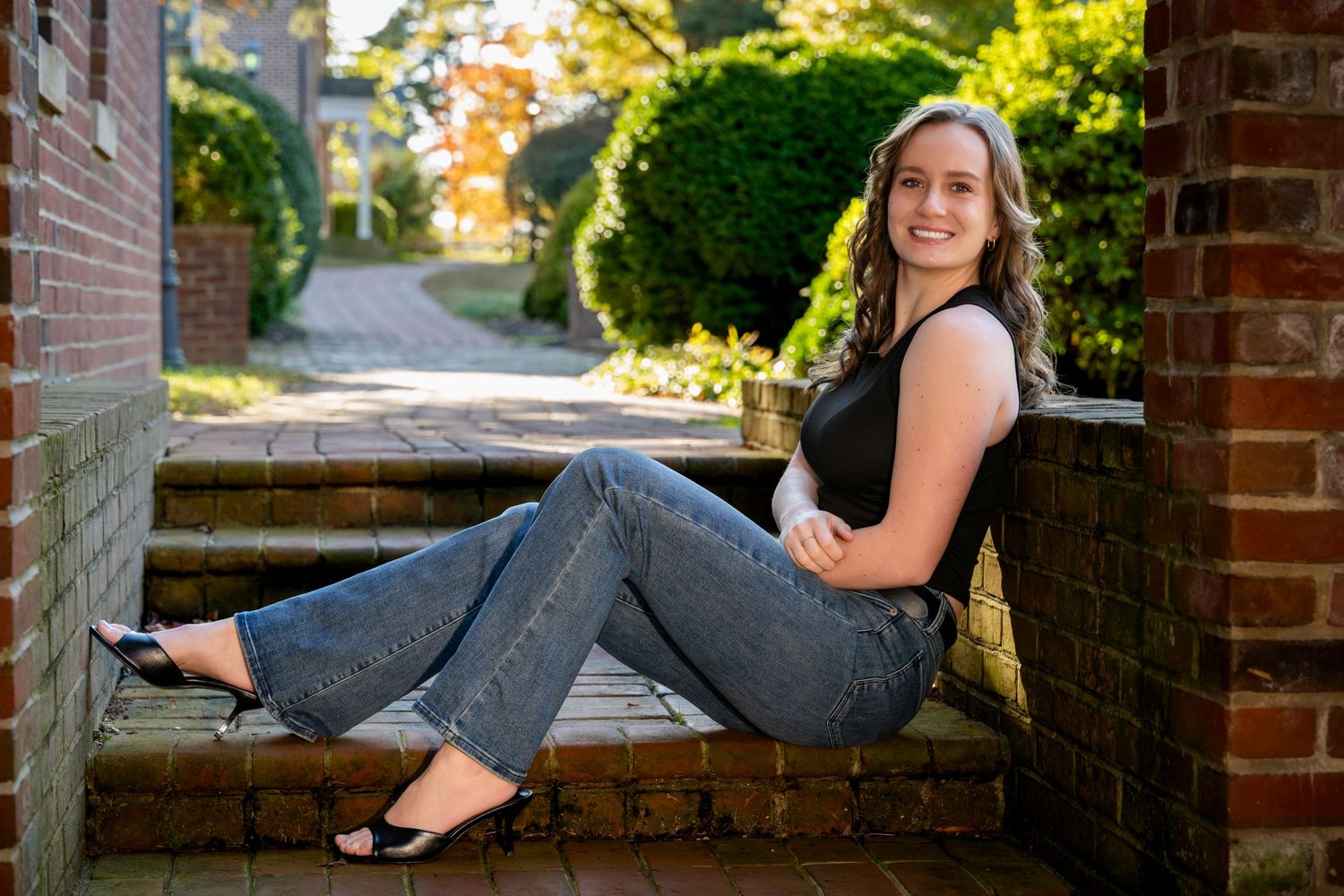 Smiling young woman in jeans and black top sitting on brick steps in a lush garden setting.
