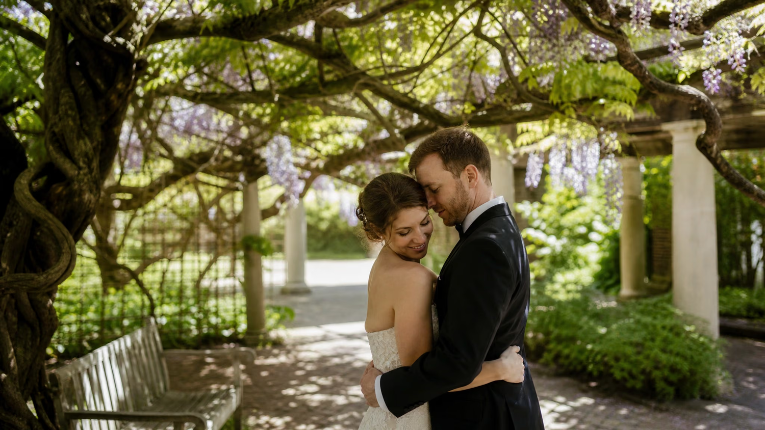Couple embracing tenderly under wisteria pergola in lush garden on their wedding day.
