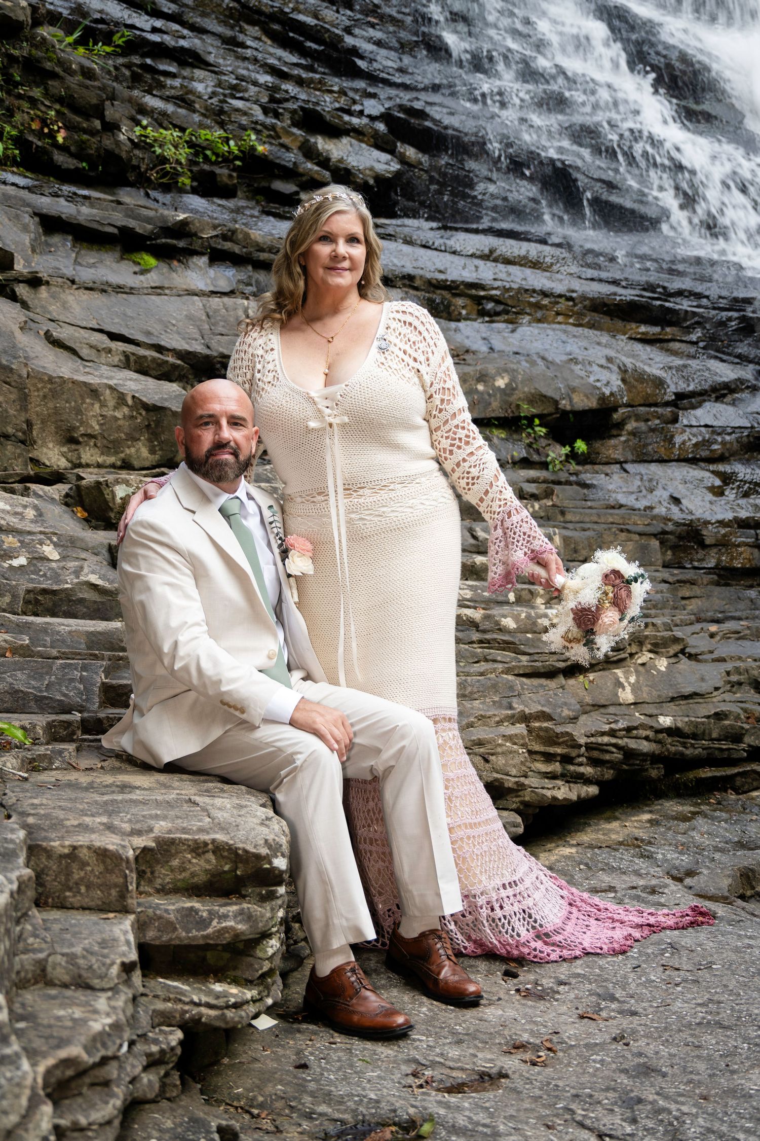 Bride and groom pose on rocky steps before a waterfall, she in a crochet gown, he in a tan suit.