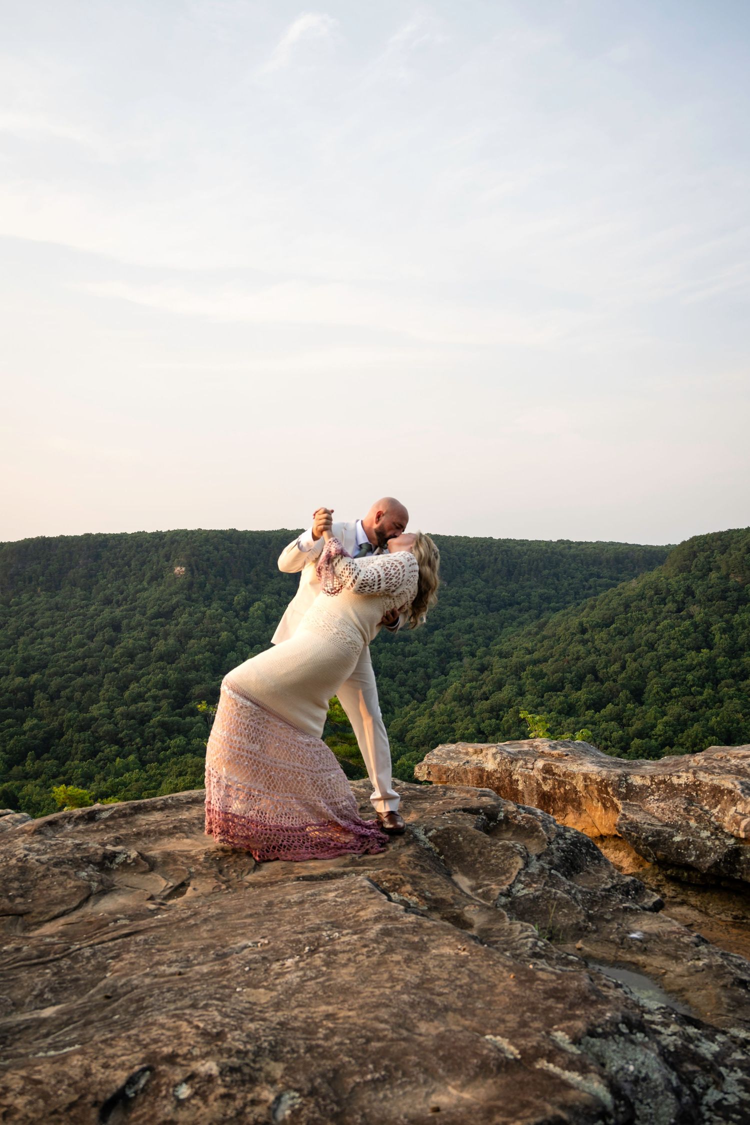 Couple sharing a romantic dip kiss on a rocky mountain overlook with lush green valley views.