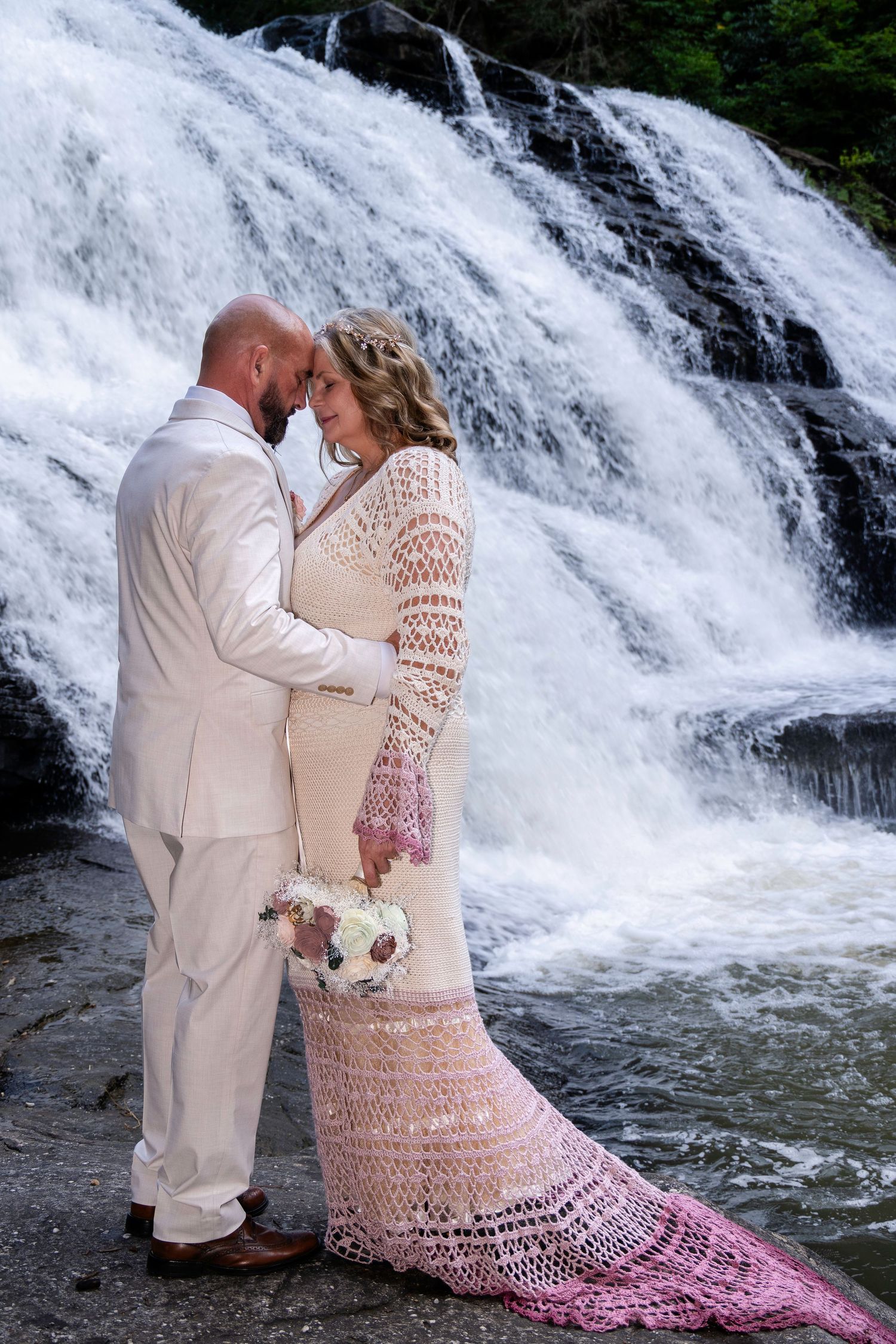 Couple sharing romantic moment in front of cascading waterfall, bride in crochet gown holding bouquet, groom in white suit.