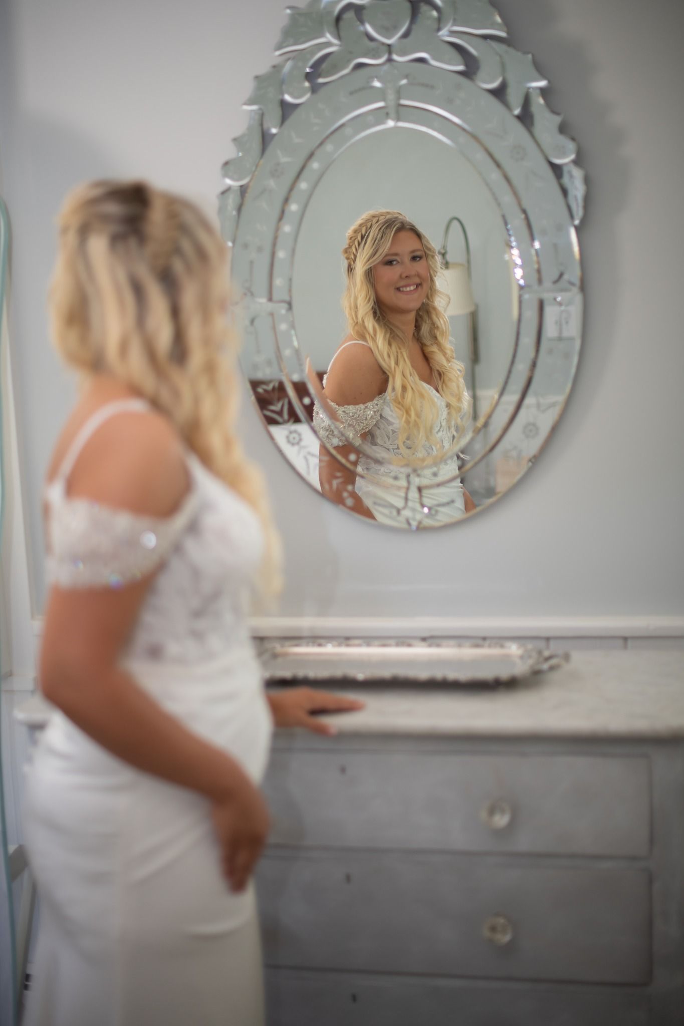 Bride in fitted white lace wedding dress admiring herself in decorative oval mirror.