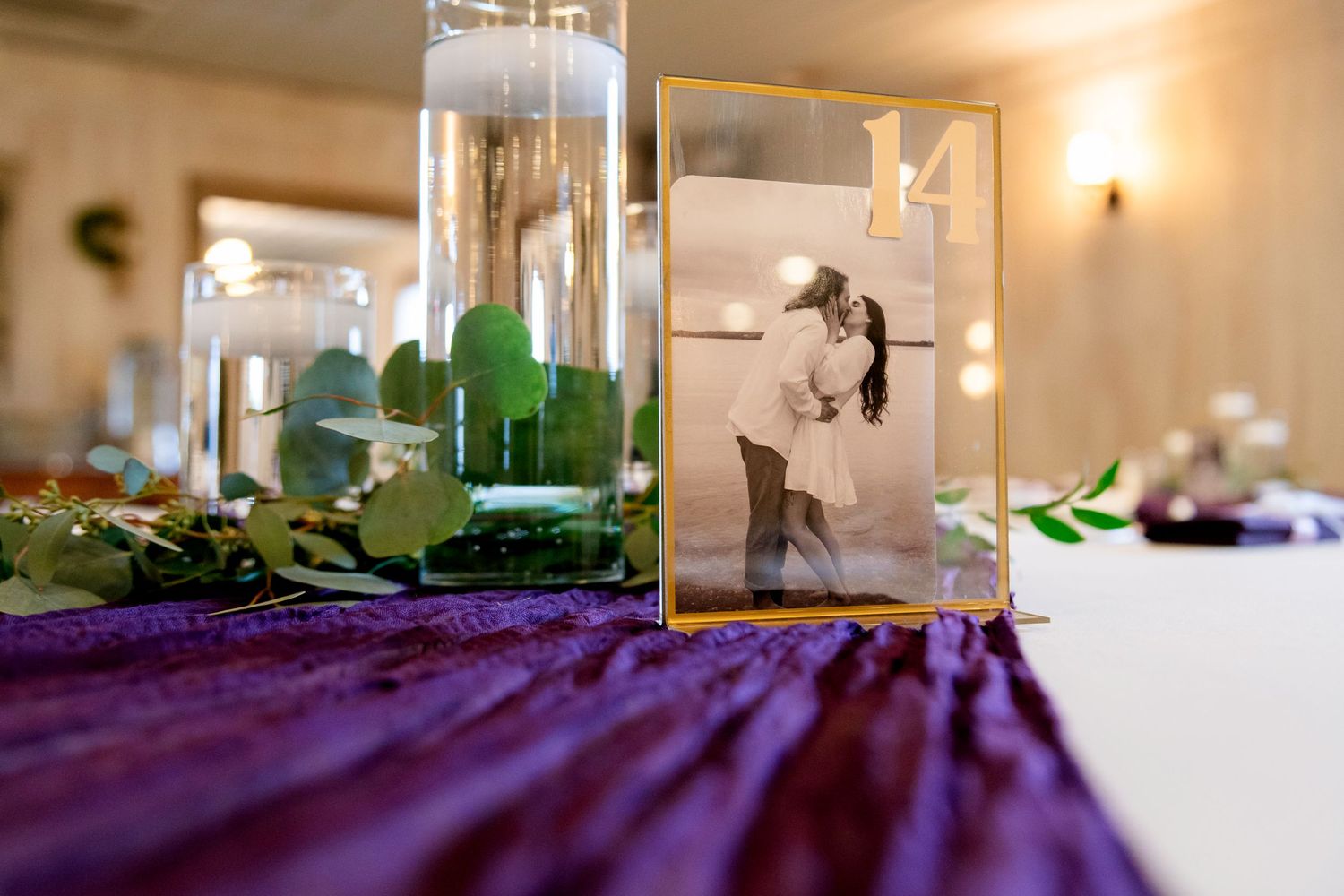 Wedding table centerpiece featuring a framed black and white couple photo, candles, and greenery on purple linen.