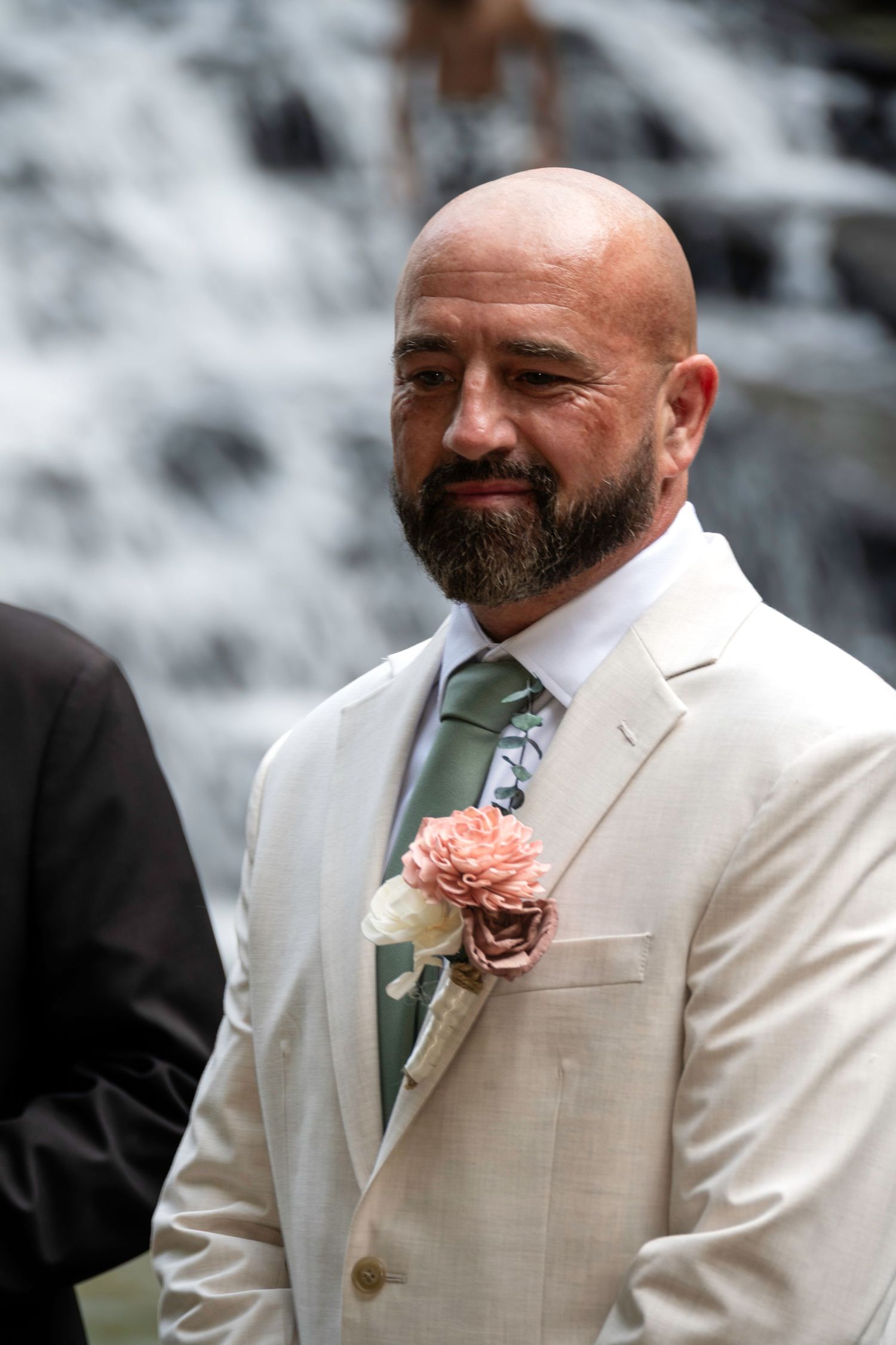 Bald groom with beard wearing cream suit, green tie, and pink boutonniere stands before a waterfall.