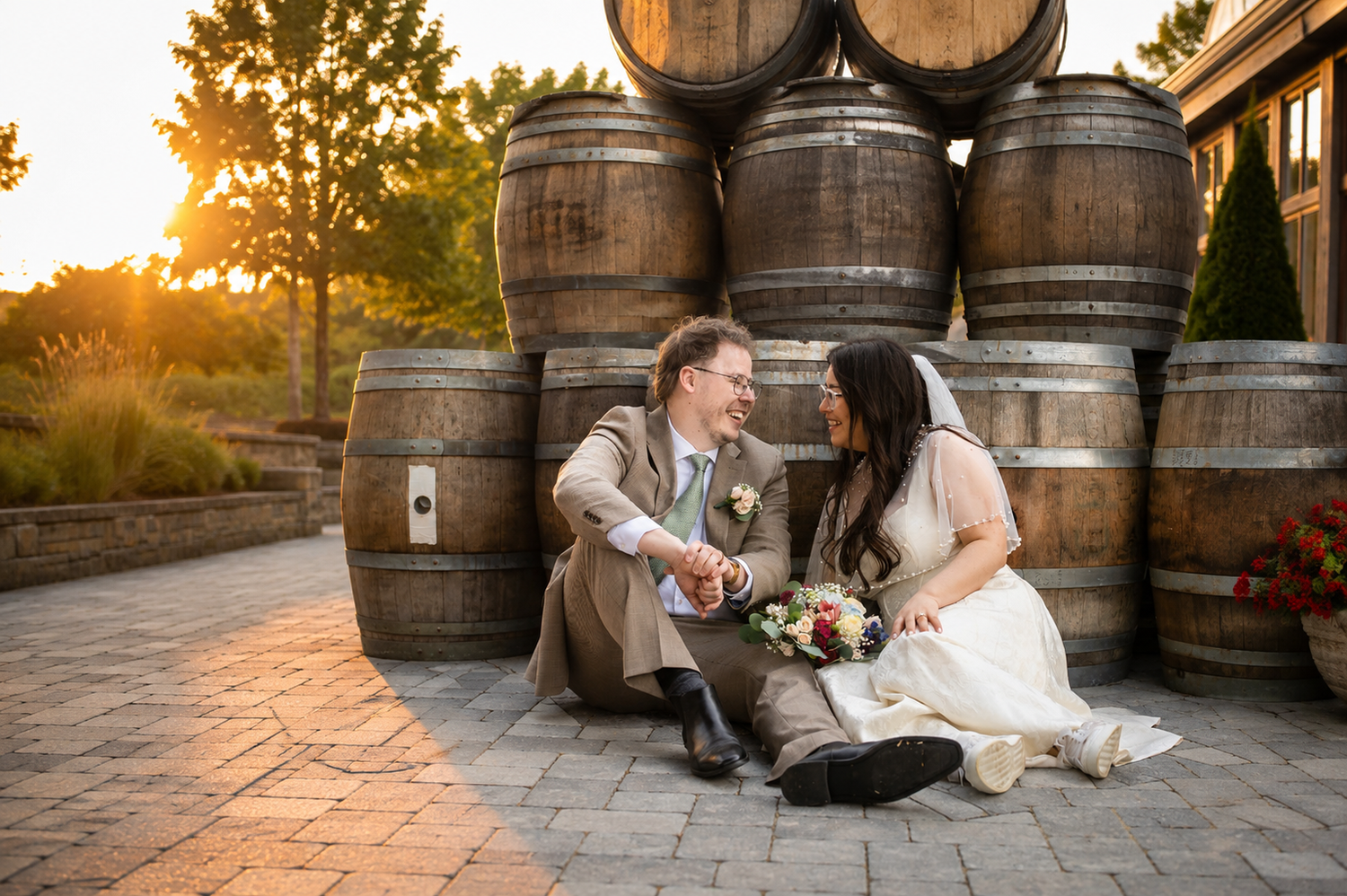 Bride and groom sitting together laughing in front of rustic wine barrels at golden hour wedding venue.