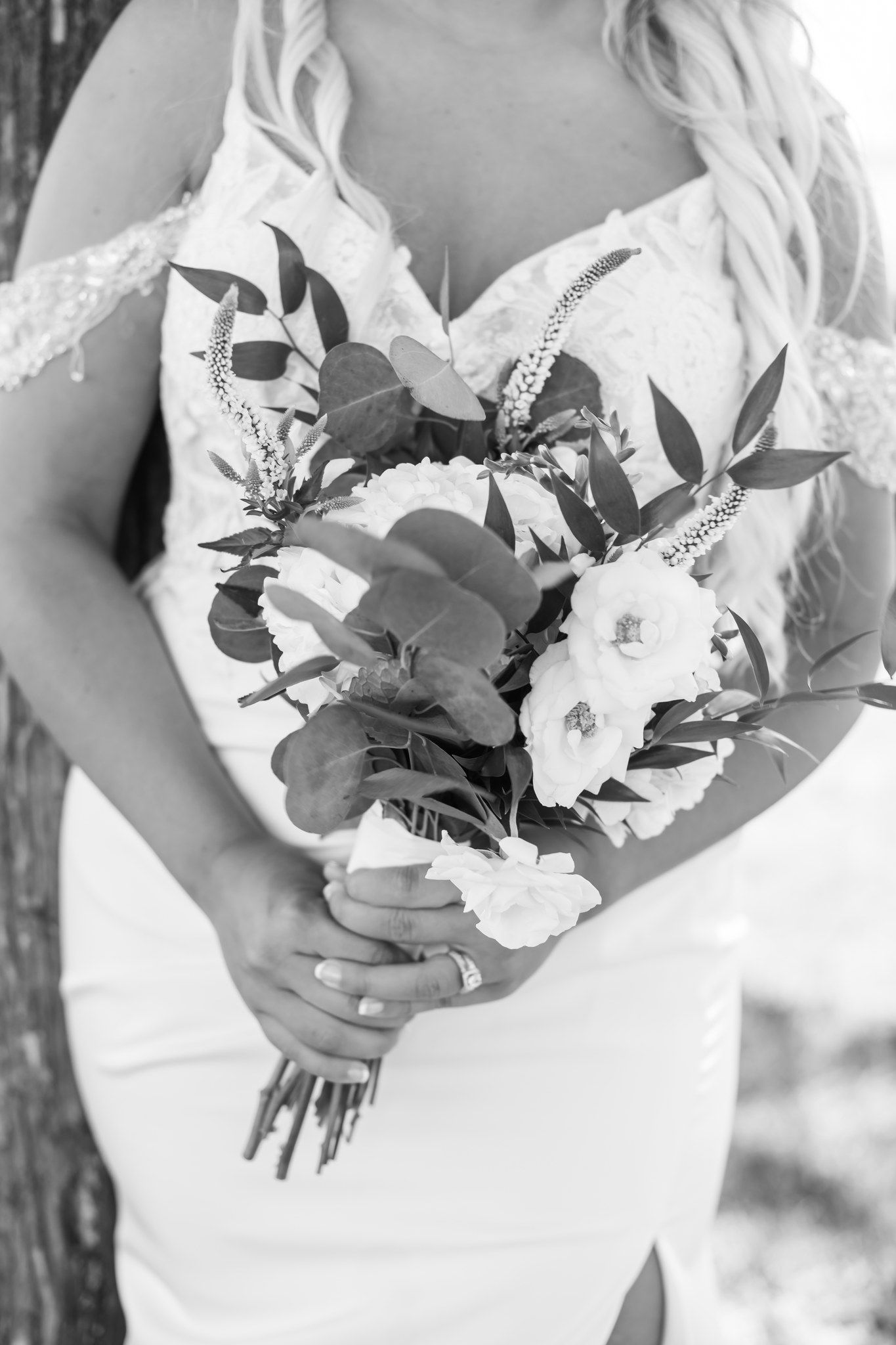 Black and white close-up of bride holding floral bouquet with greenery in lace gown.