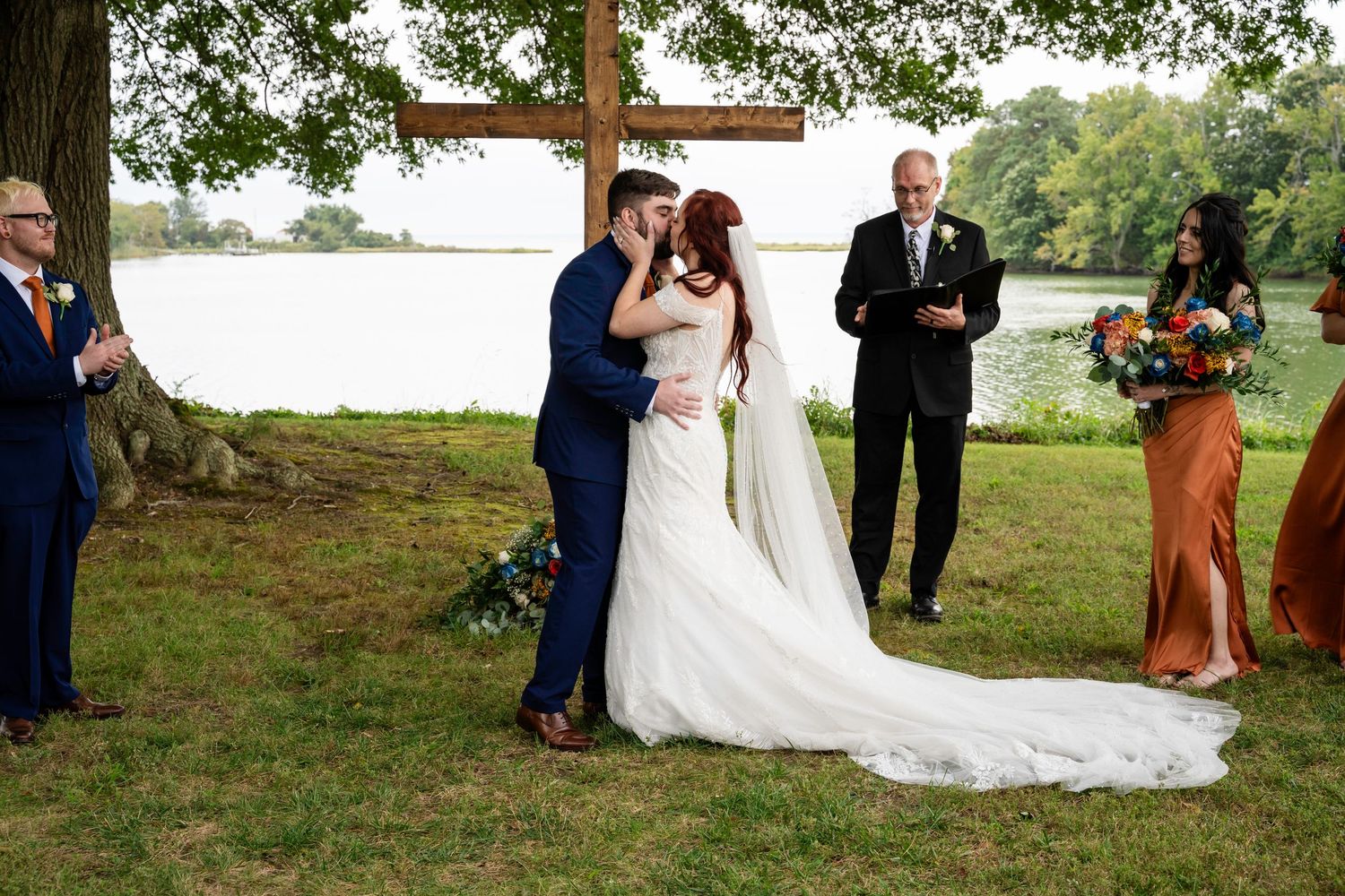 A newlywed couple shares their first kiss under a large tree with a wooden cross by a peaceful lake.
