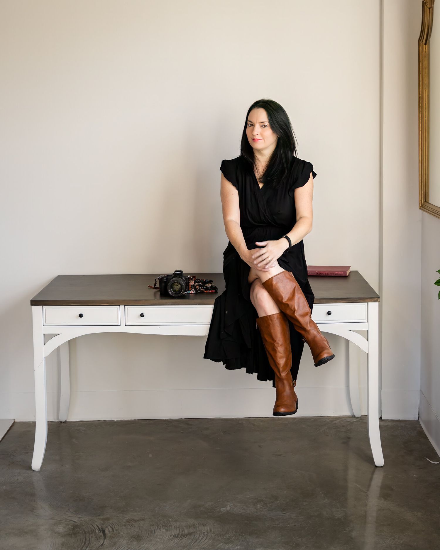 Female photographer in black dress and brown boots sitting on white desk with camera in minimalist studio.