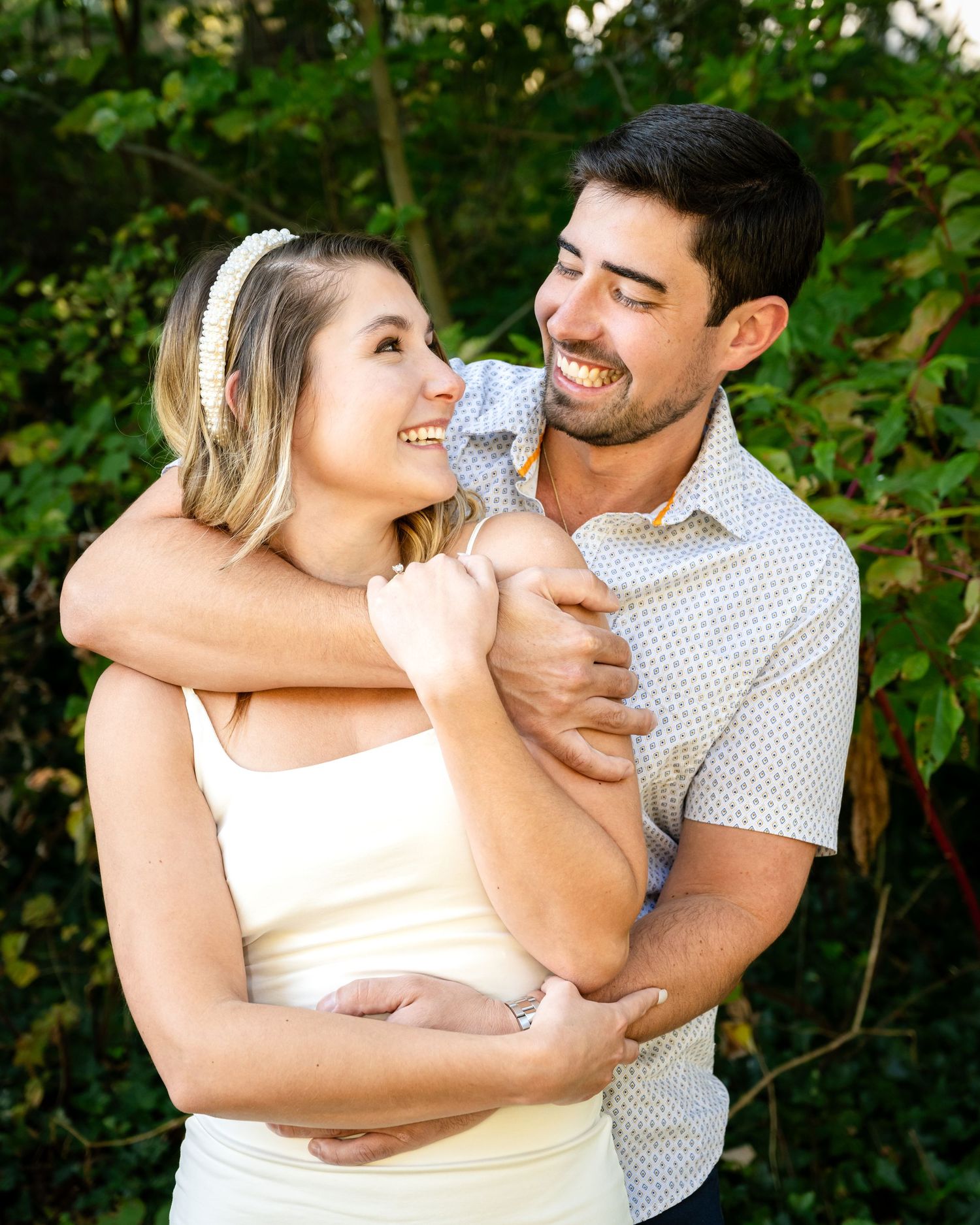 A smiling couple embraces outdoors, with the man hugging the woman from behind among lush green foliage.