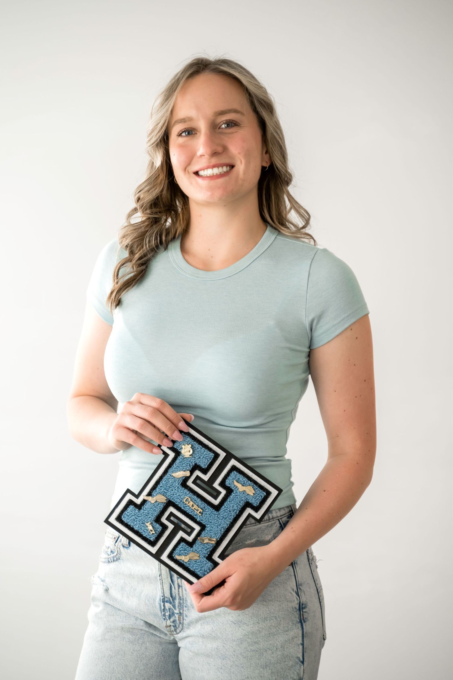 Smiling young woman in mint green t-shirt holding a blue varsity letter H patch against white background.
