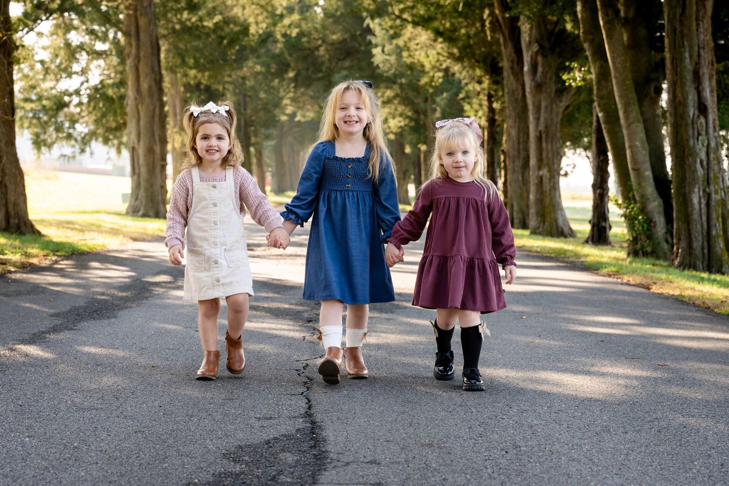 Three young girls in dresses hold hands walking down a tree-lined path outdoors.