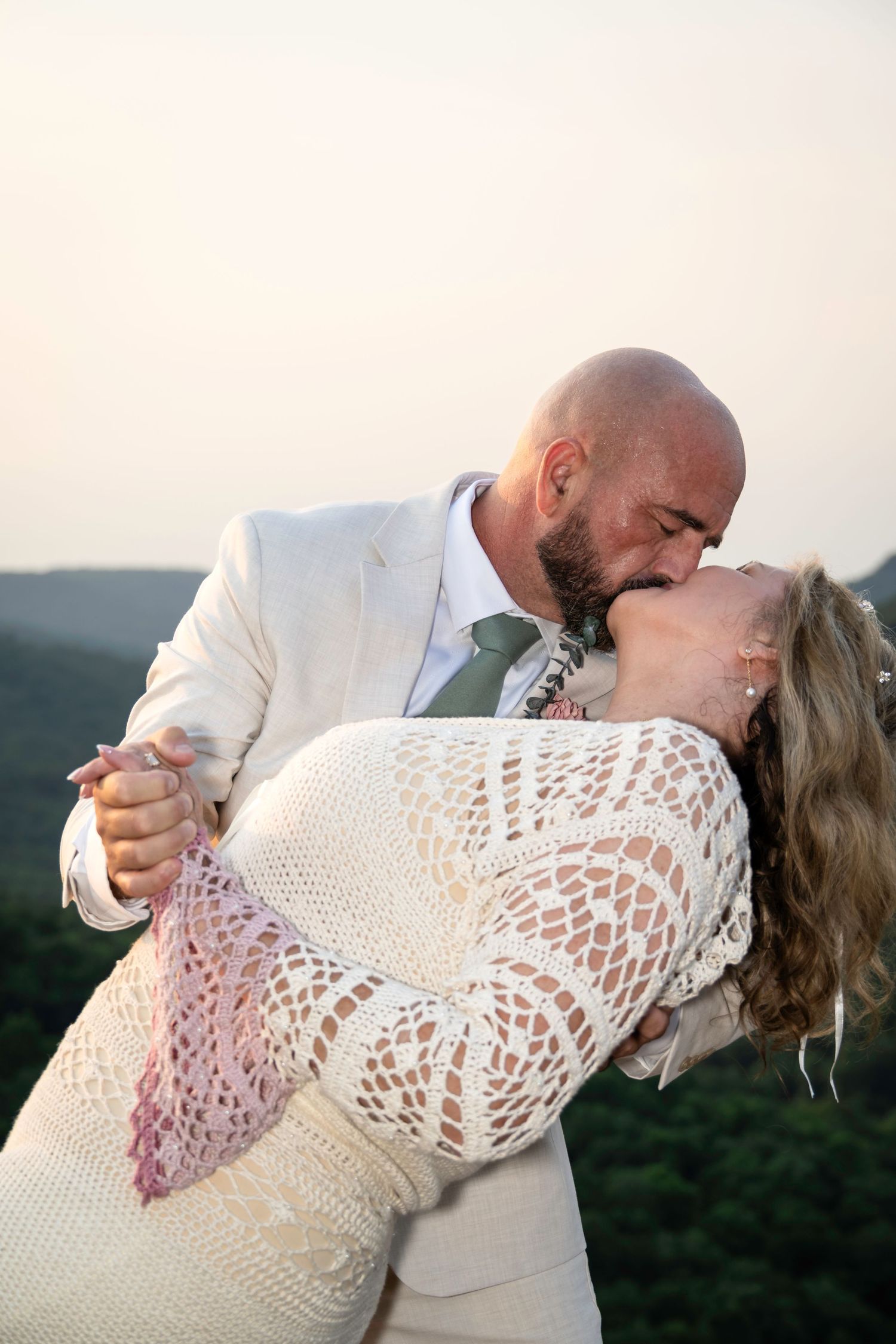 Bride and groom share a romantic dip kiss outdoors at sunset, with lush green hills in the background.
