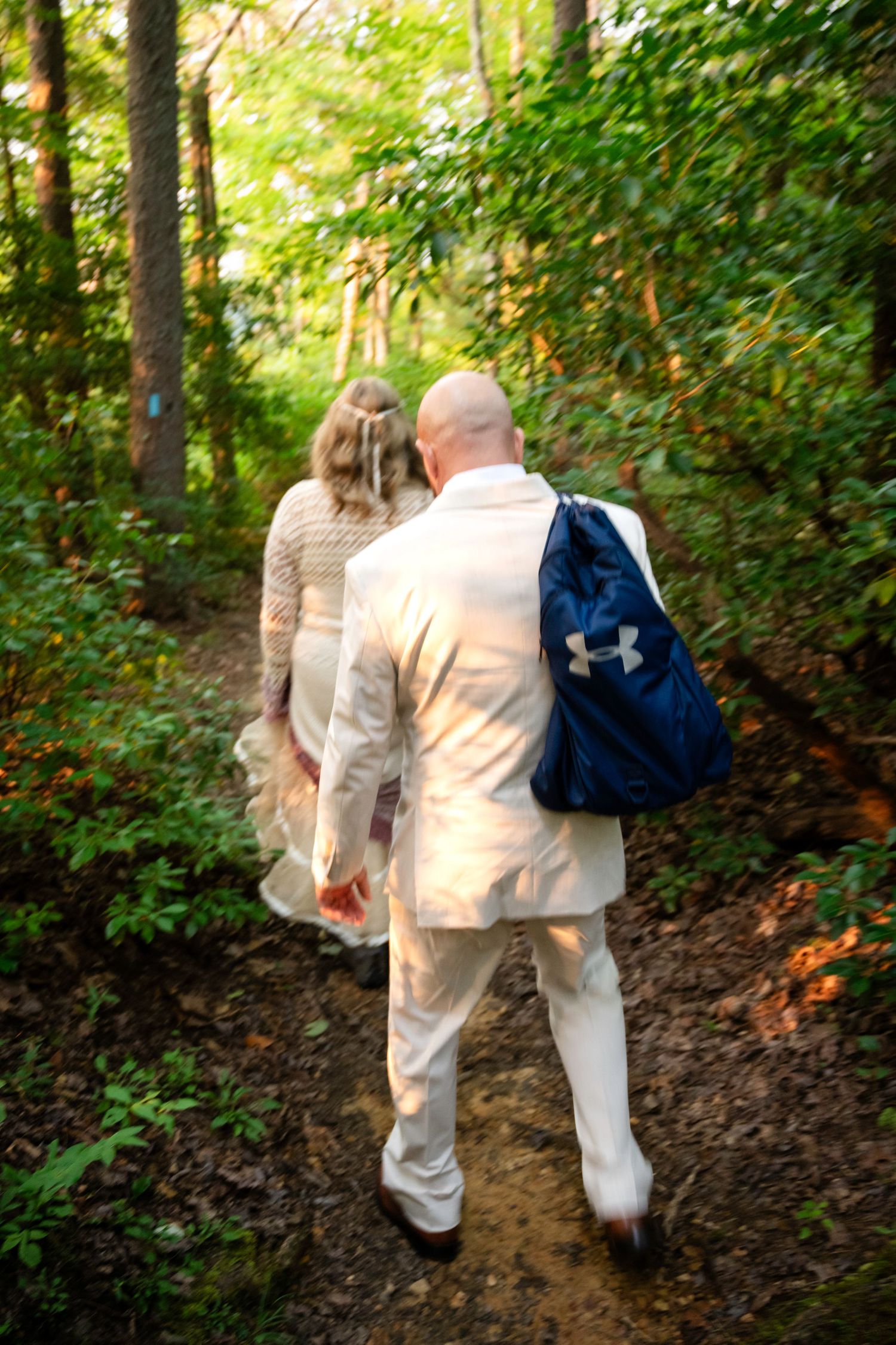 A bald groom in a white suit with a blue Under Armour backpack walks with a bride along a forest trail.