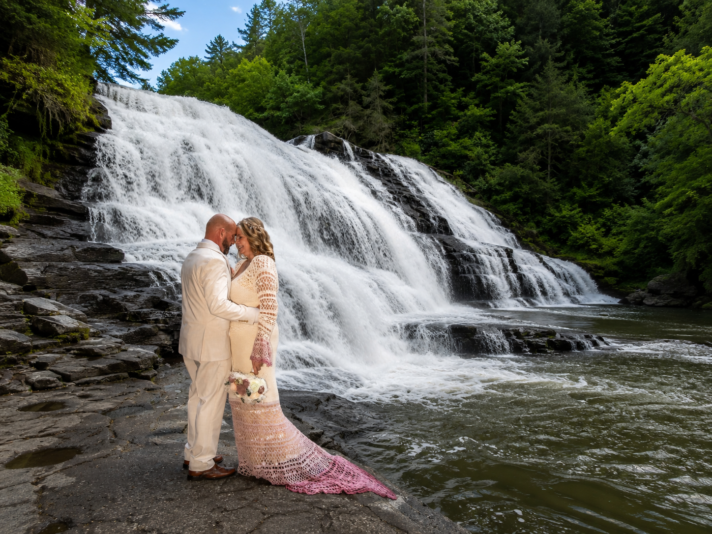 Couple kissing in wedding attire before a stunning cascading waterfall surrounded by lush green forest.