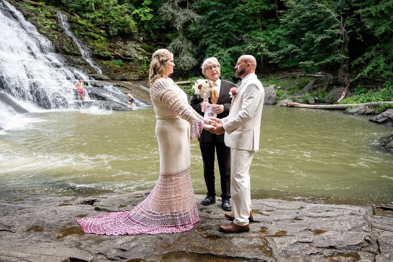 Couple exchanges vows with officiant beside a scenic waterfall on rocky terrain during an outdoor wedding ceremony.