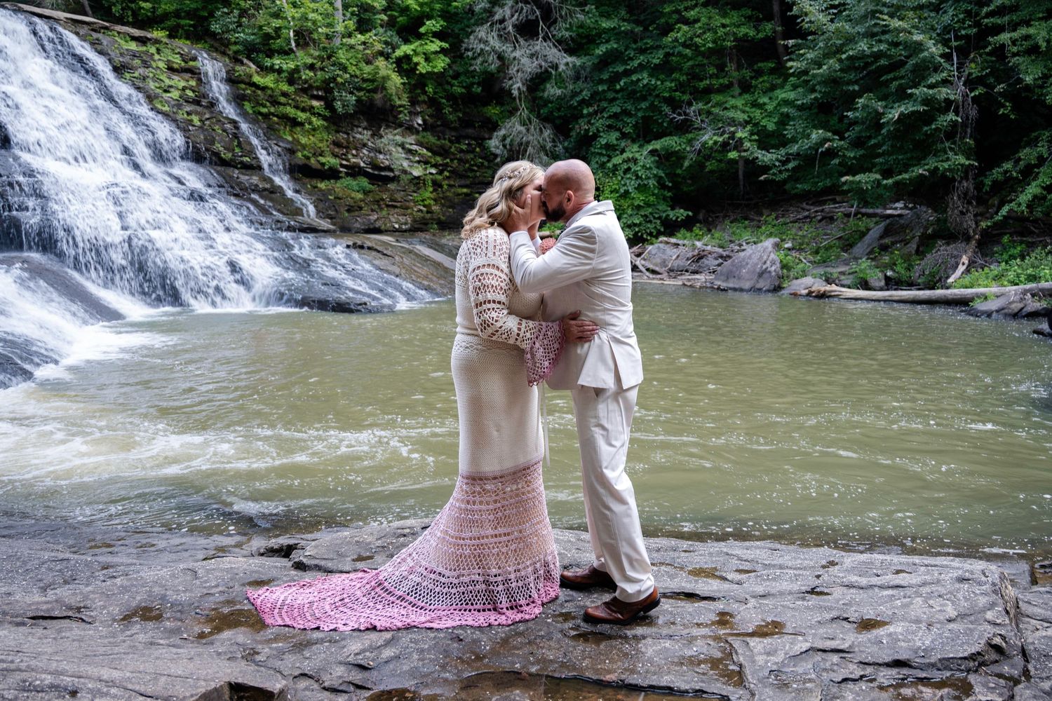 Couple sharing a romantic kiss in wedding attire beside a scenic waterfall on rocky terrain surrounded by lush greenery.