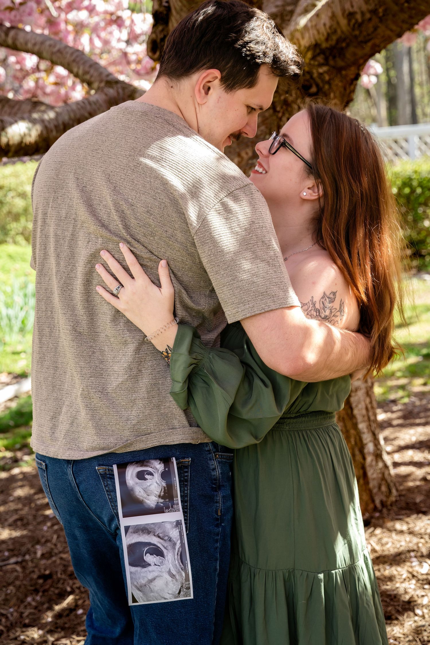 Couple embracing under cherry blossom tree holding ultrasound photos, pregnancy announcement outdoors.