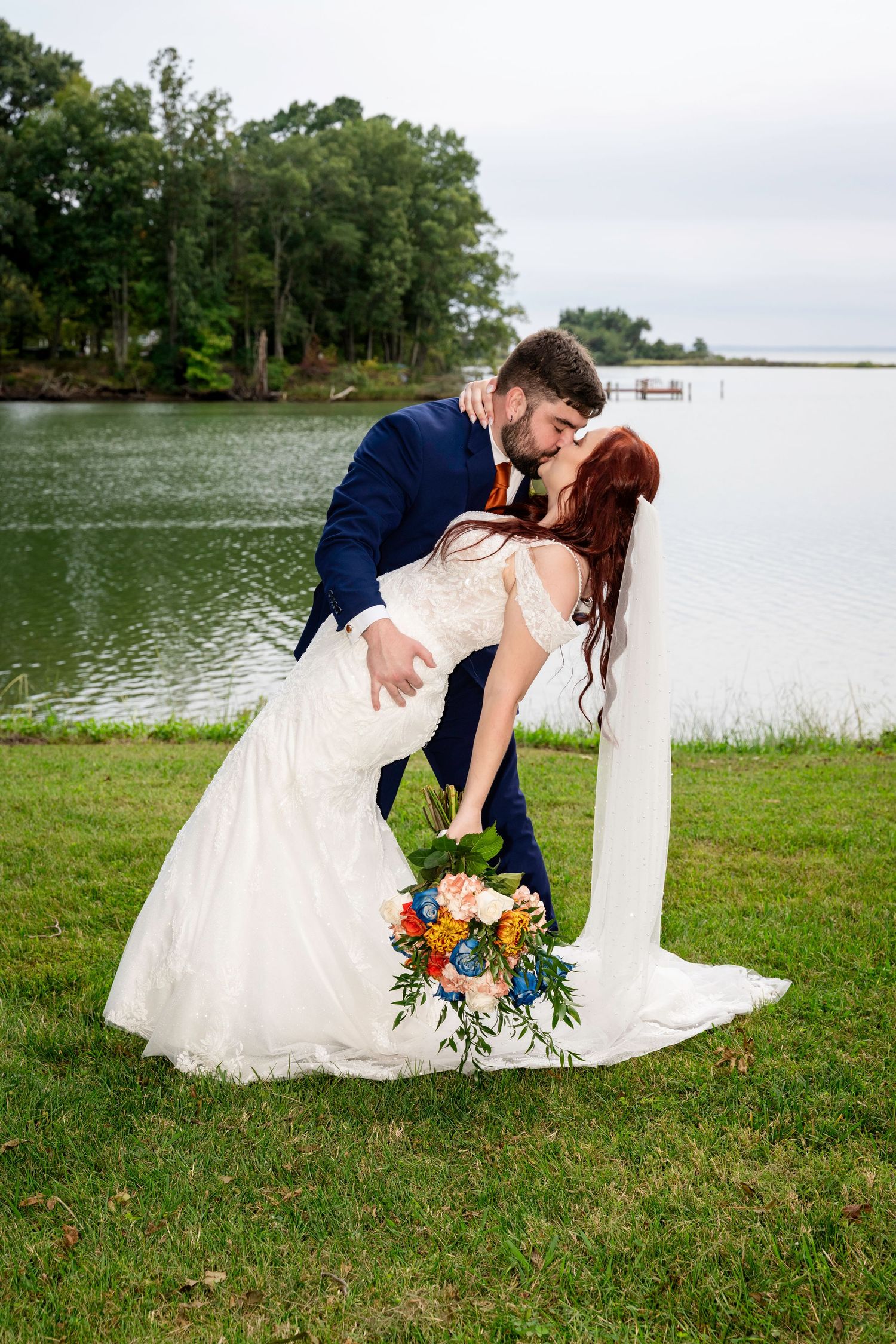 Groom dips bride for a romantic kiss by a lake holding a colorful bouquet.