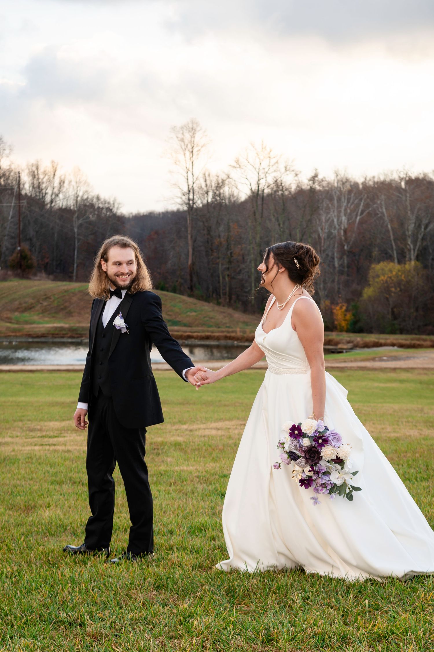Bride and groom hold hands in a green field with autumn trees and pond backdrop on their wedding day.