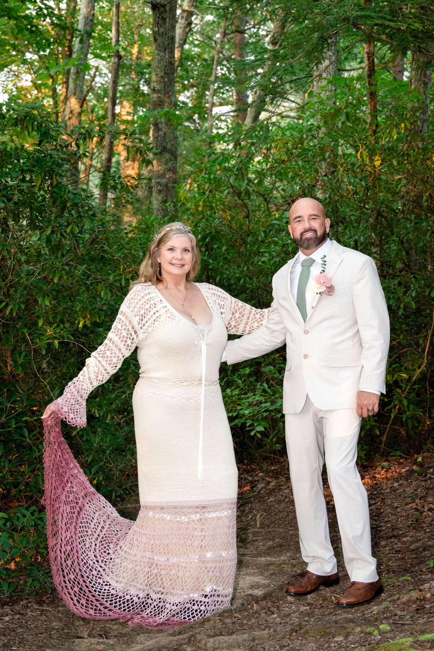 Bride in white crochet gown holding pink shawl stands with groom in cream suit outdoors in lush green forest setting.
