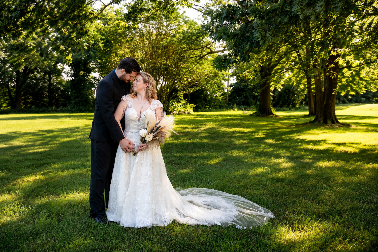 Bride and groom share a romantic moment under lush green trees on a sunny wedding day.