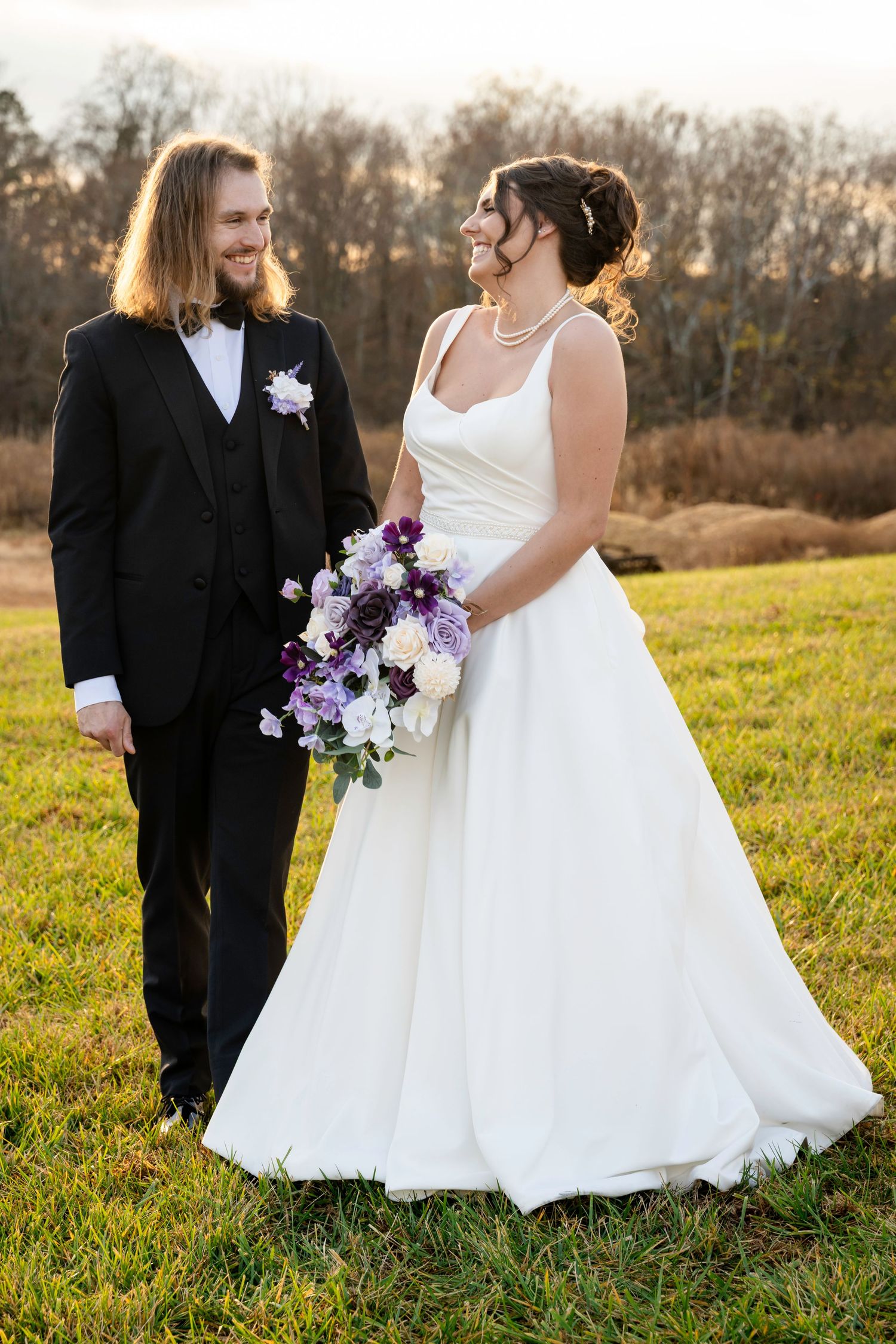 Bride holds purple and white bouquet while couple gazes at each other in outdoor field.
