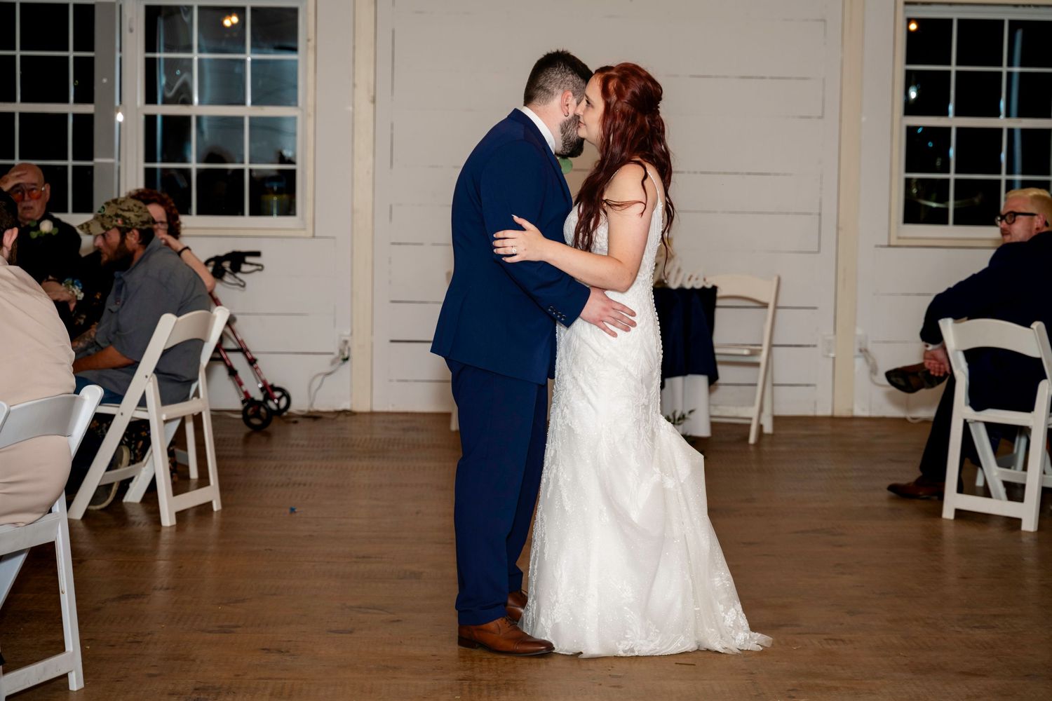 Newlyweds share an intimate first dance in a white-walled reception hall while guests watch.