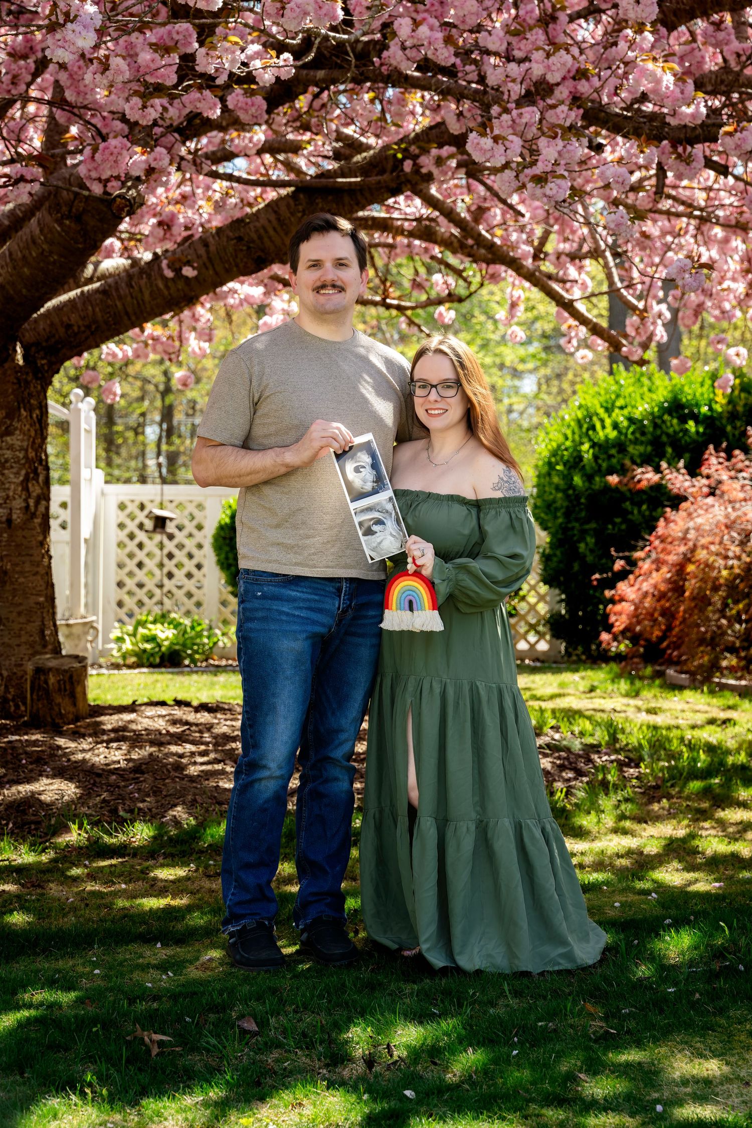 Couple holding ultrasound photos and rainbow sign under pink cherry blossom tree for pregnancy announcement.