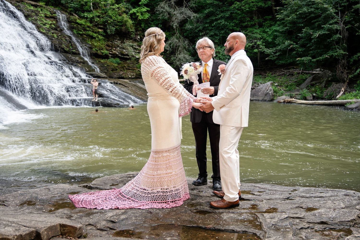 Couple exchanging vows at a waterfall wedding ceremony on rocky terrain, officiant holding flowers, lush green forest backdrop.